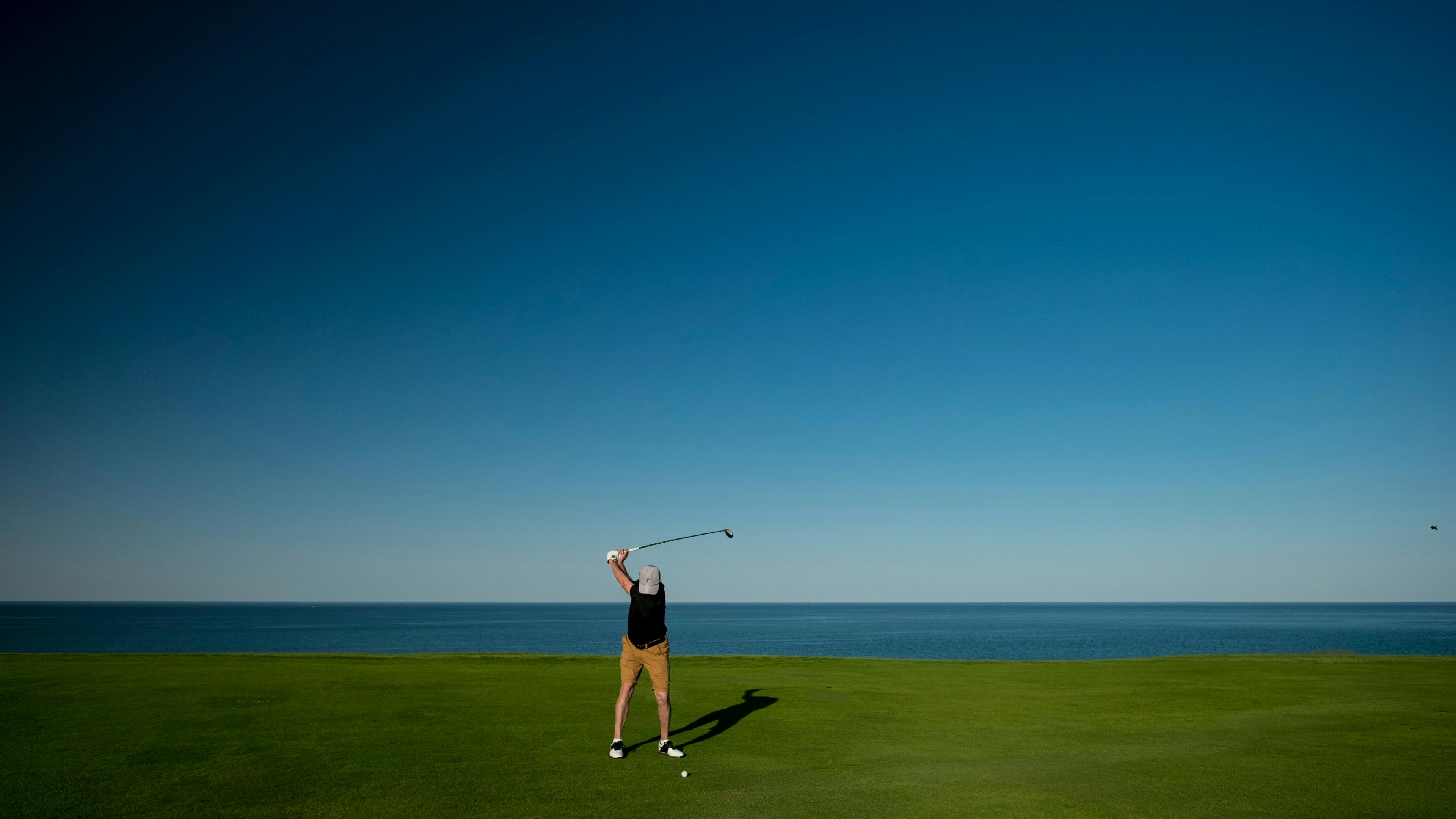 A golfer, viewed from behind, is in mid-swing on a green golf course. The expansive blue ocean and sky form a striking backdrop, with a distinct horizon line.


