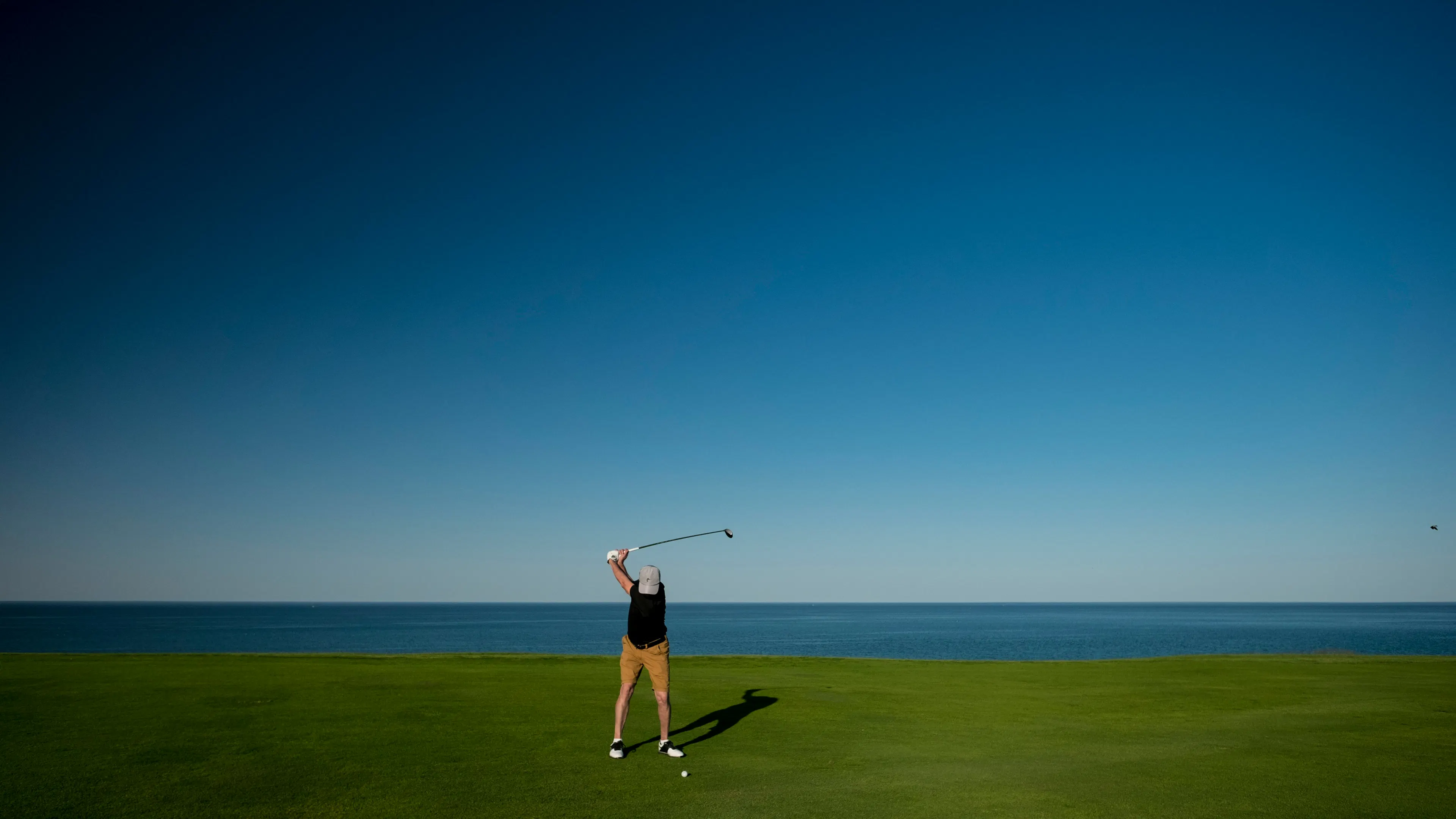 A golfer, viewed from behind, is in mid-swing on a green golf course. The expansive blue ocean and sky form a striking backdrop, with a distinct horizon line.
