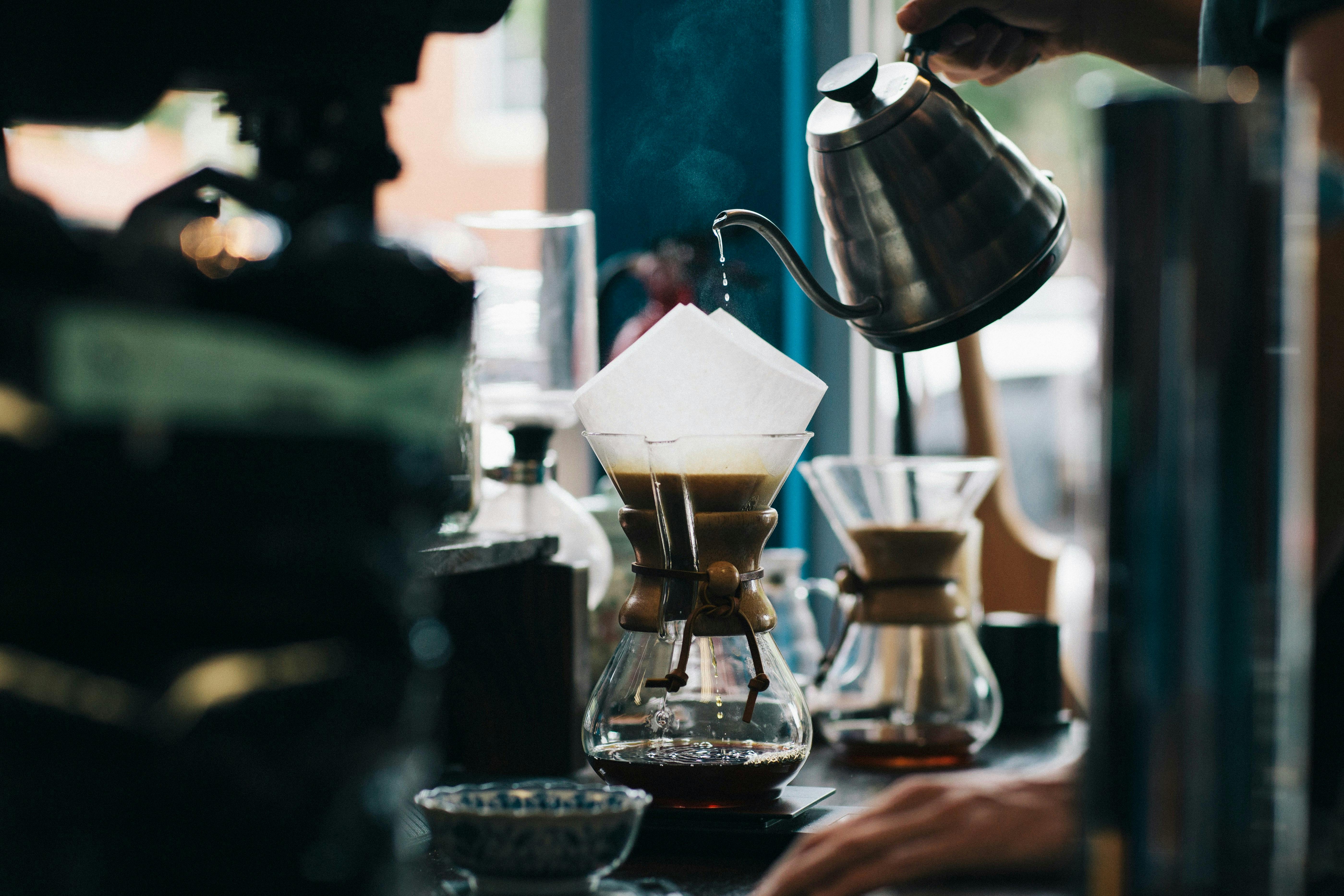 A person's hand holds a silver gooseneck kettle, pouring hot water into a Chemex coffee maker. The brew drips down into the glass carafe below. Steam rises from the top, creating a soft, ethereal effect. The background is a dimly lit coffee shop counter.