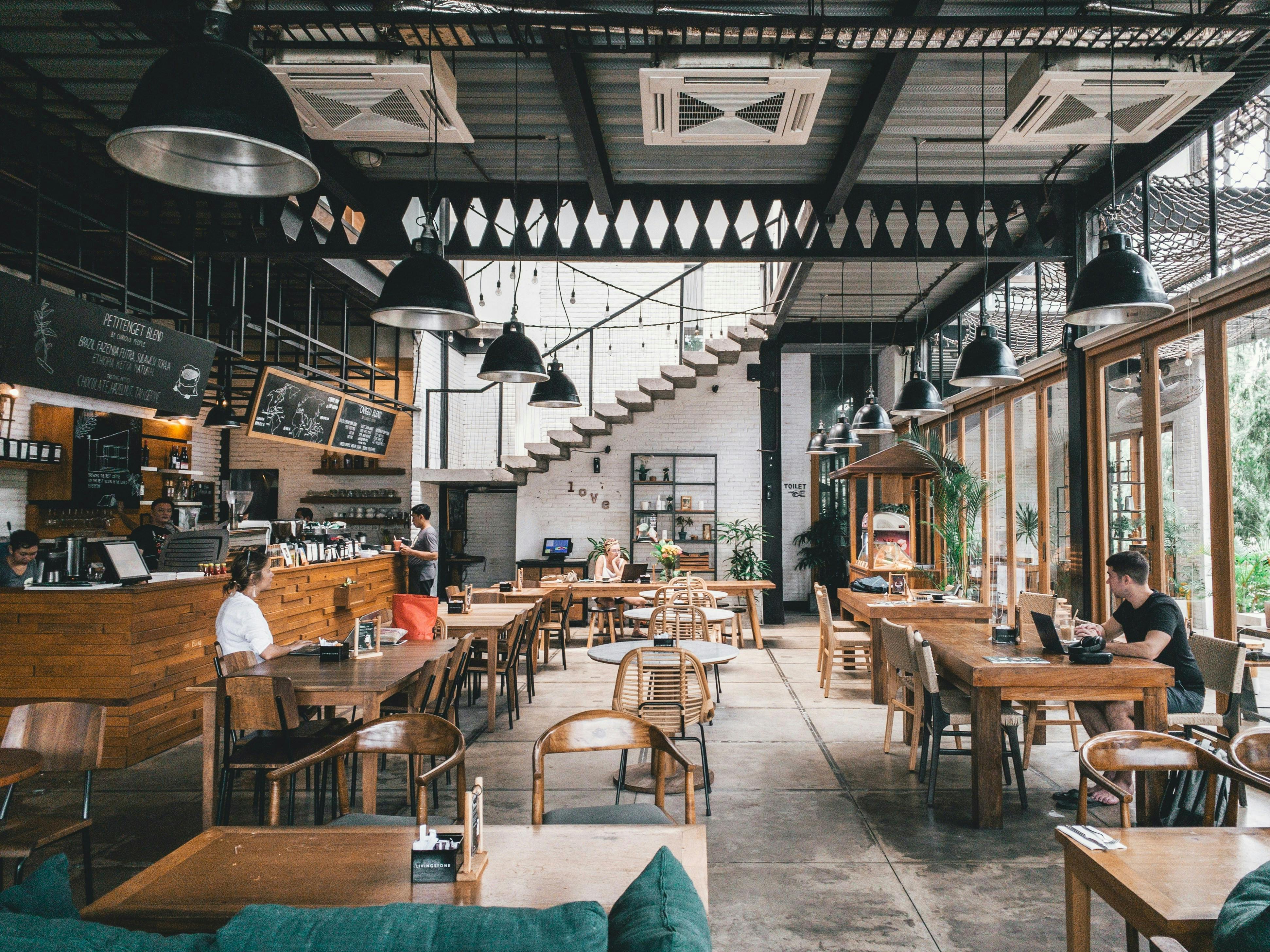 A spacious, modern coffee shop with high ceilings and large windows. The interior features a mix of communal wooden tables, wicker chairs, and a long counter. Patrons are seated at various tables, some working on laptops. A white staircase with a black railing leads to a second floor.