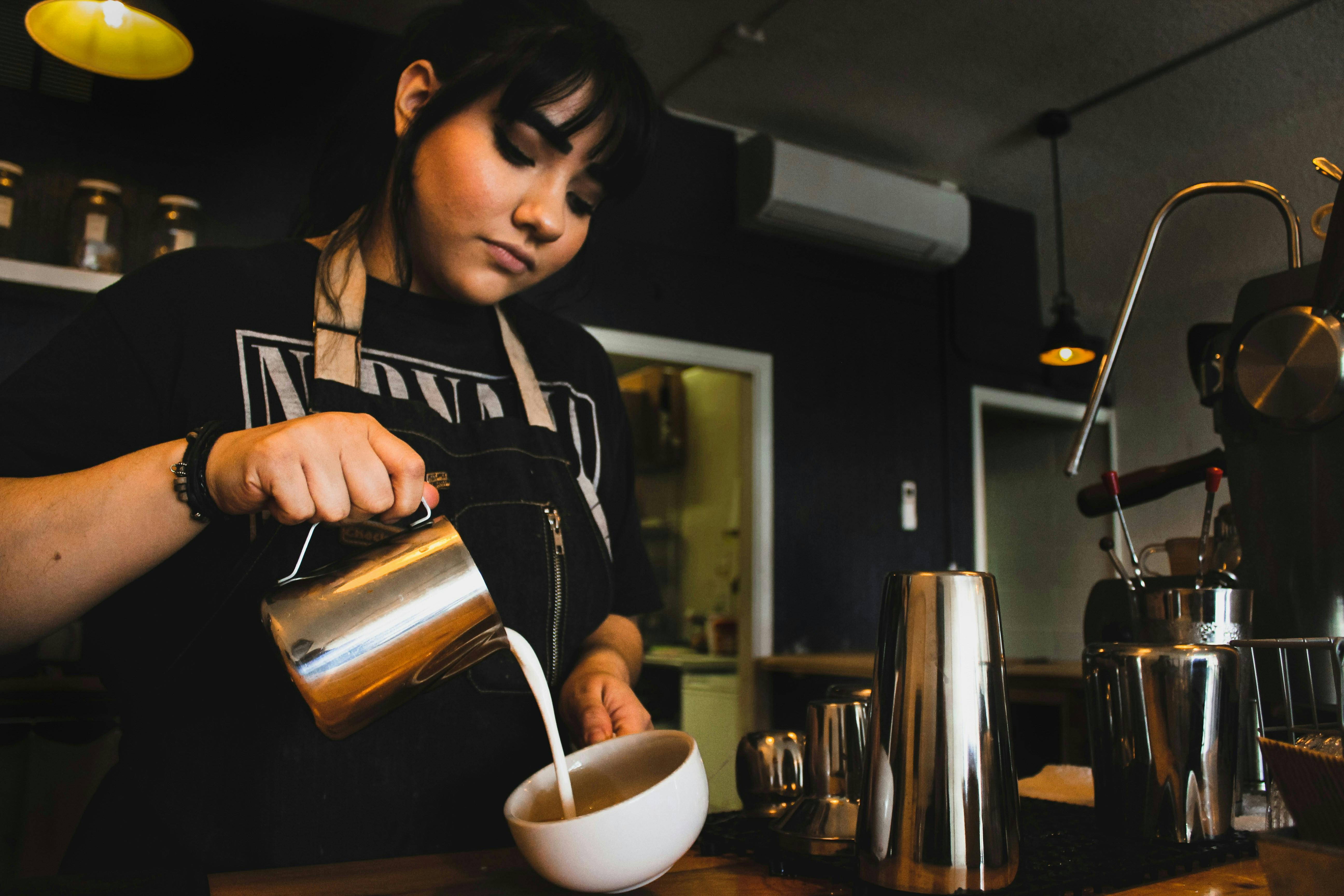 A female barista with dark hair and a Nirvana t-shirt is focused on pouring frothed milk from a silver pitcher into a white coffee cup. The scene is shot from a low angle, highlighting her concentration on creating latte art.

