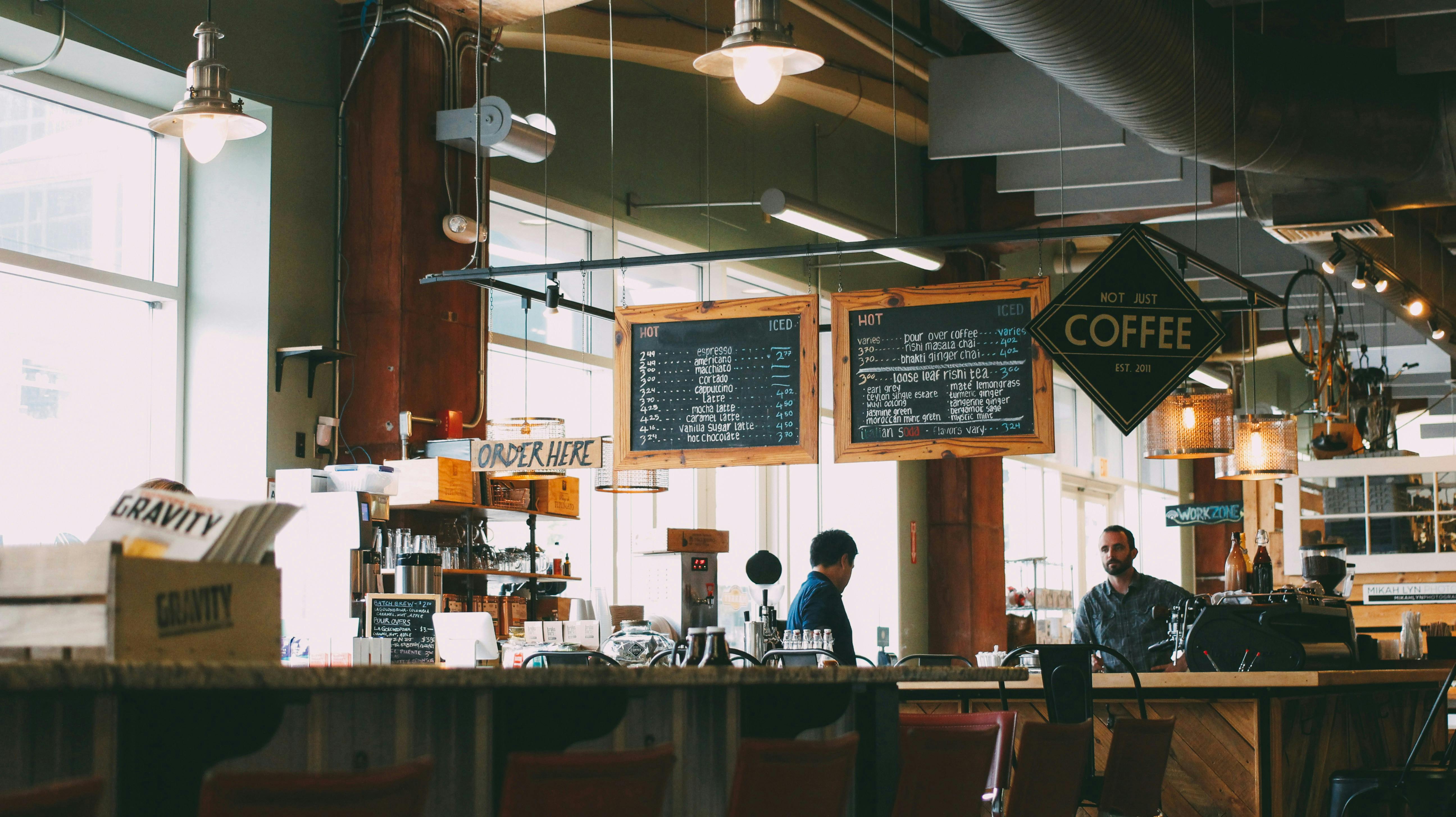 The interior of a casual coffee shop with a focus on the counter and menu boards. Two chalkboards hang from the ceiling, listing "ICED" and "HOT" drink options. A sign that reads "NOT JUST COFFEE" is also visible. Two people are standing behind the counter, one appears to be taking an order.