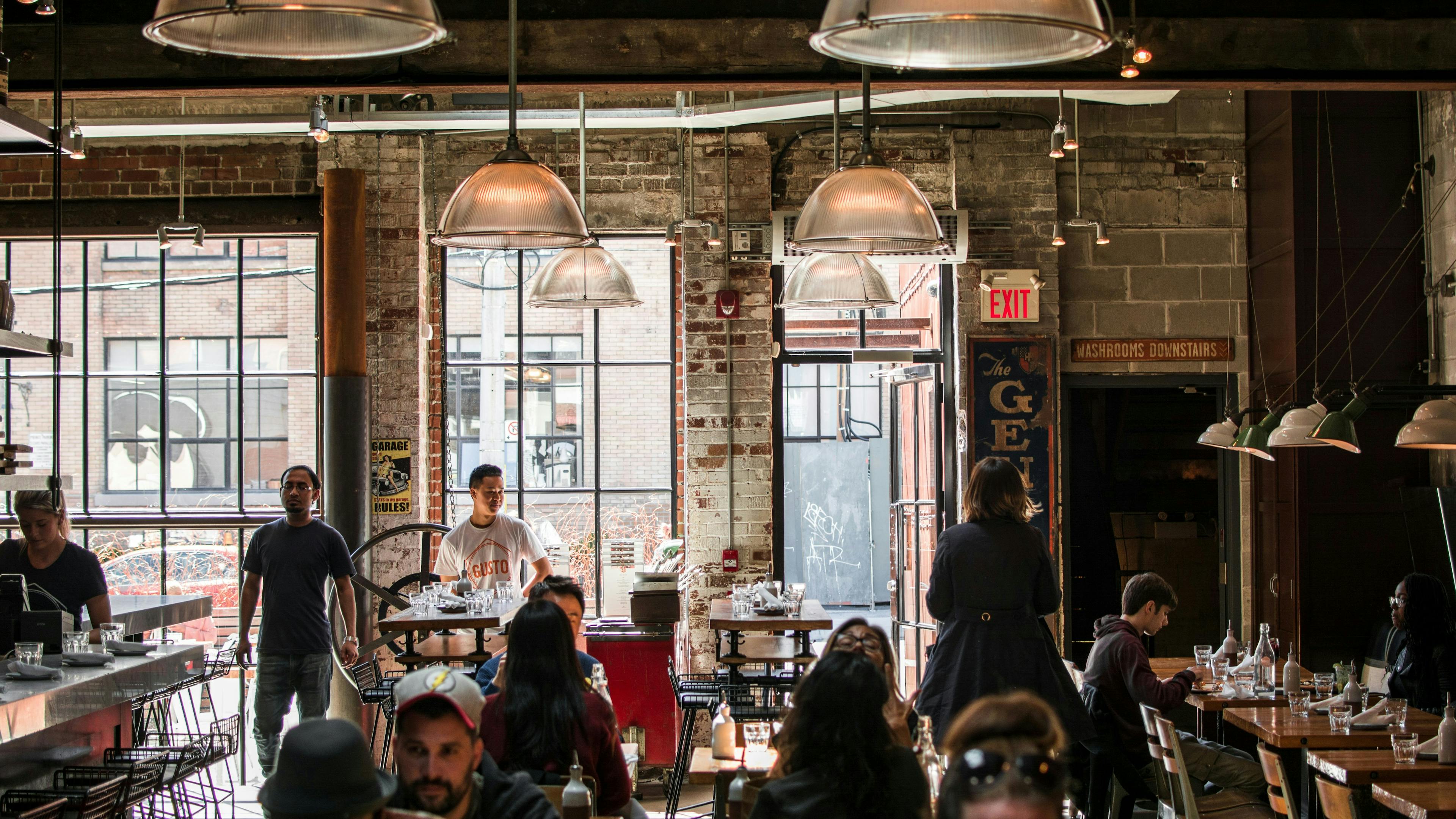 A lively, crowded coffee shop with an industrial-style interior. People are seated at tables and a bar counter, some talking and others on their phones. The walls are exposed brick and large windows look out onto a city street. Large, metallic pendant lights hang from the ceiling.