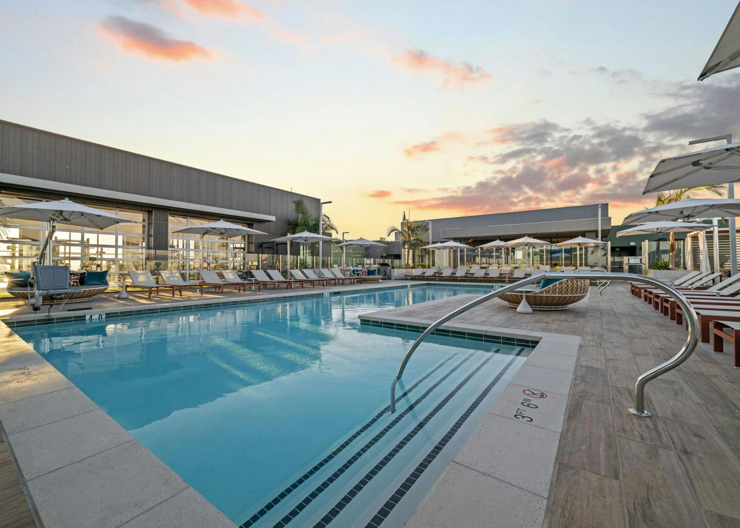 Rooftop pool deck at sunset featuring a modern swimming pool, lounge chairs, umbrellas, and sleek architecture in the background.