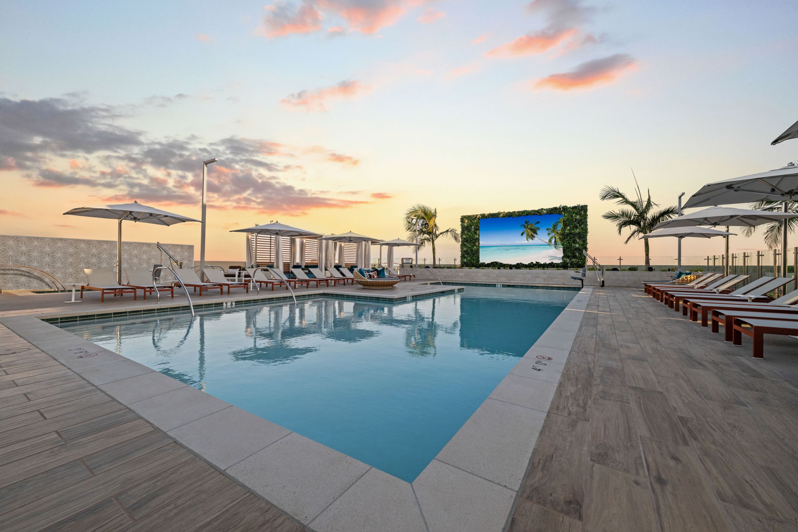 Dusk view of sparkling blue pool at AMLI Aero with recliners, large tv, fitness center building and palm trees and panoramic rooftop.