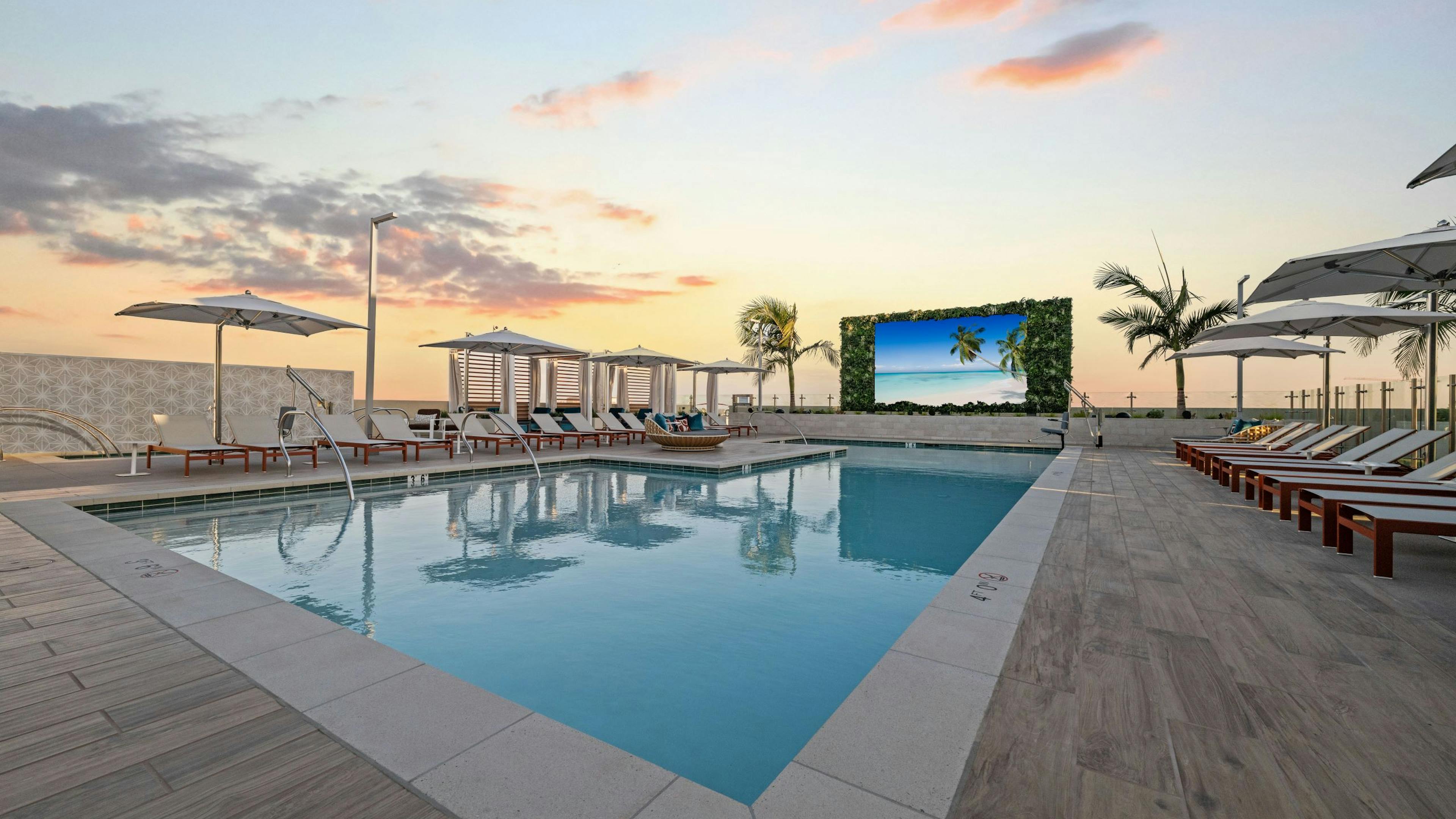 Dusk view of sparkling blue pool at AMLI Aero with recliners, large tv, fitness center building and palm trees and panoramic rooftop.