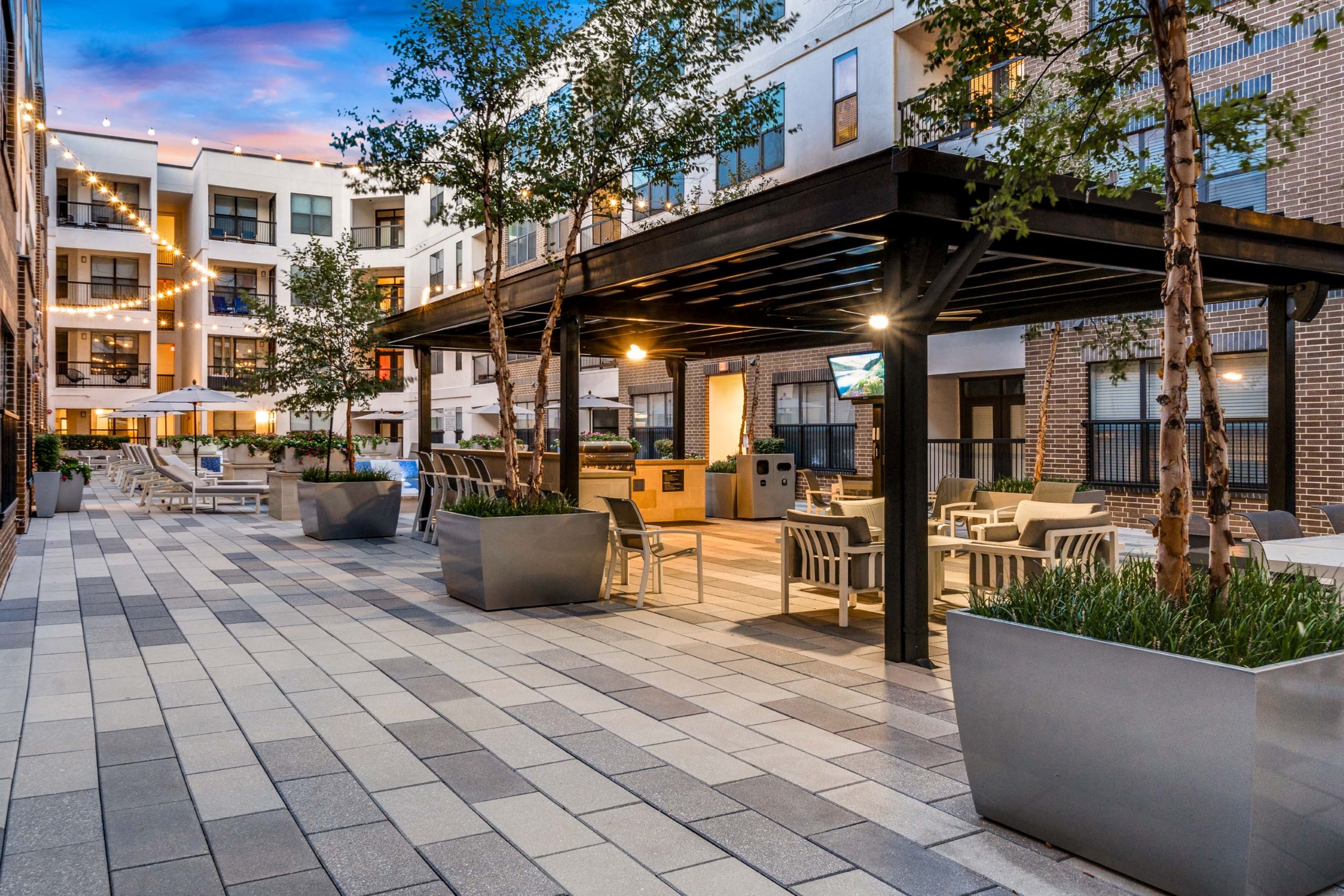 AMLI Quadrangle covered barbeque grill and seating area with television and surrounding planted trees with string lights in the background.