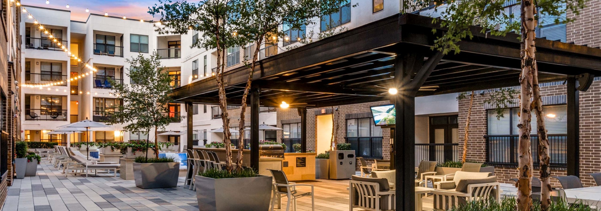 AMLI Quadrangle covered barbeque grill and seating area with television and surrounding planted trees with string lights in the background.