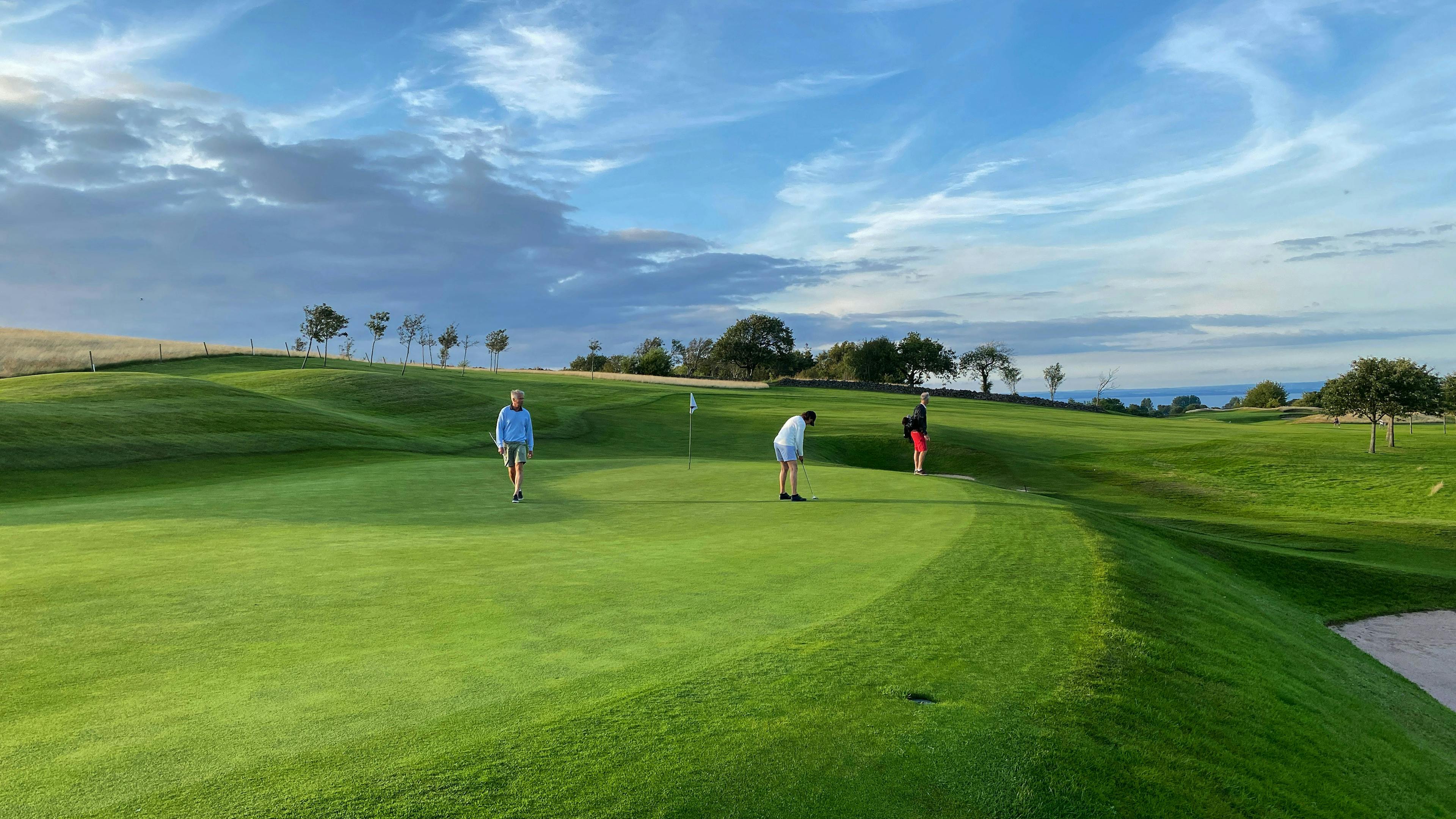 Three people are playing golf on a vast, undulating green course under a dramatic, cloudy blue sky at sunset. The golfer in the center is bent over, lining up a putt near the hole flag.