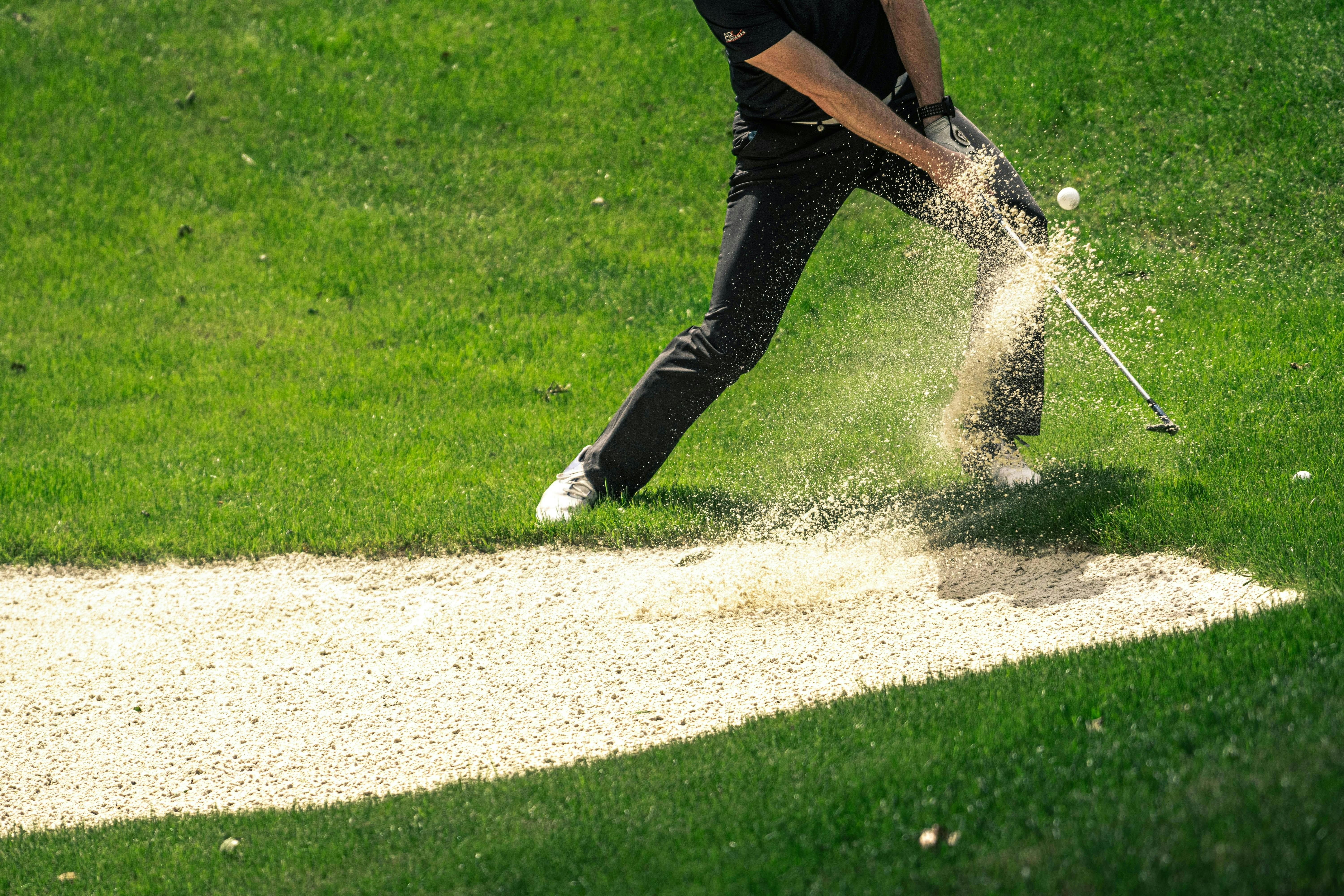 A golfer, wearing black pants and a dark shirt, hits a golf ball from a patch of sand next to a grassy area, causing a spray of sand and dirt. Only the lower body of the golfer and the action of the sand explosion are visible.