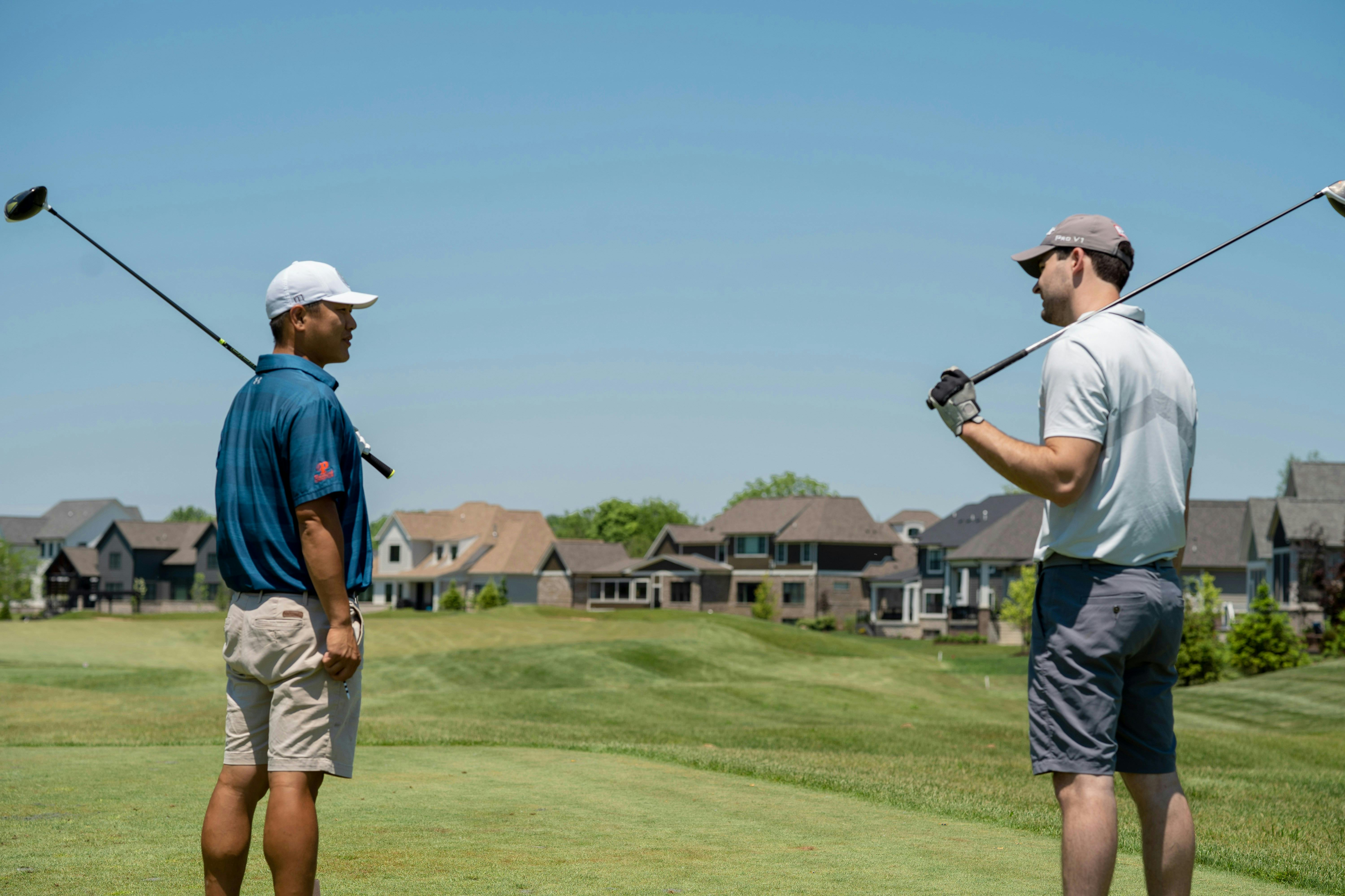 Two male golfers, one in a blue shirt and white hat, the other in a light grey shirt and dark hat, stand on a sunny green fairway talking to each other. They both have drivers resting on their shoulders. A suburban neighborhood of houses is visible in the background.