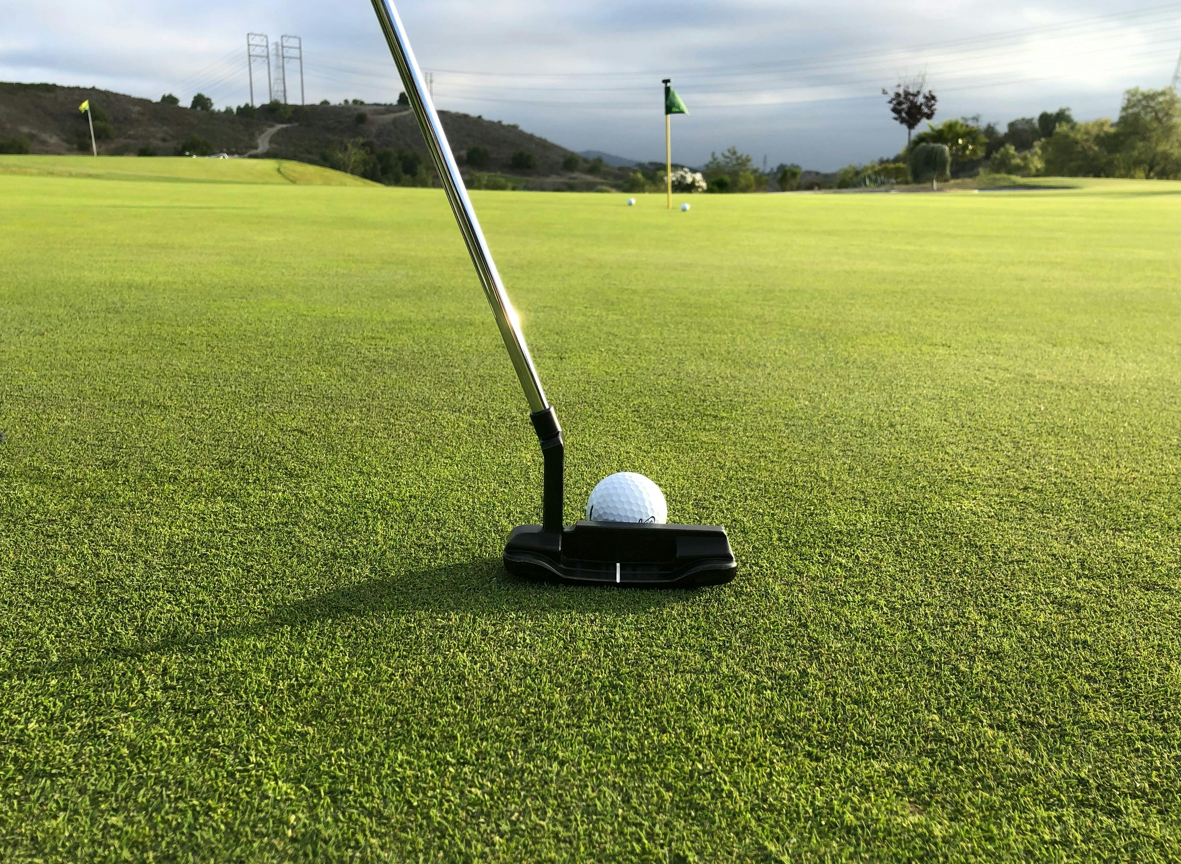 A low-angle, close-up shot of a putter head about to strike a golf ball on a meticulously manicured putting green. A flagstick is visible in the distance, and the background shows rolling hills and power lines under a cloudy sky.