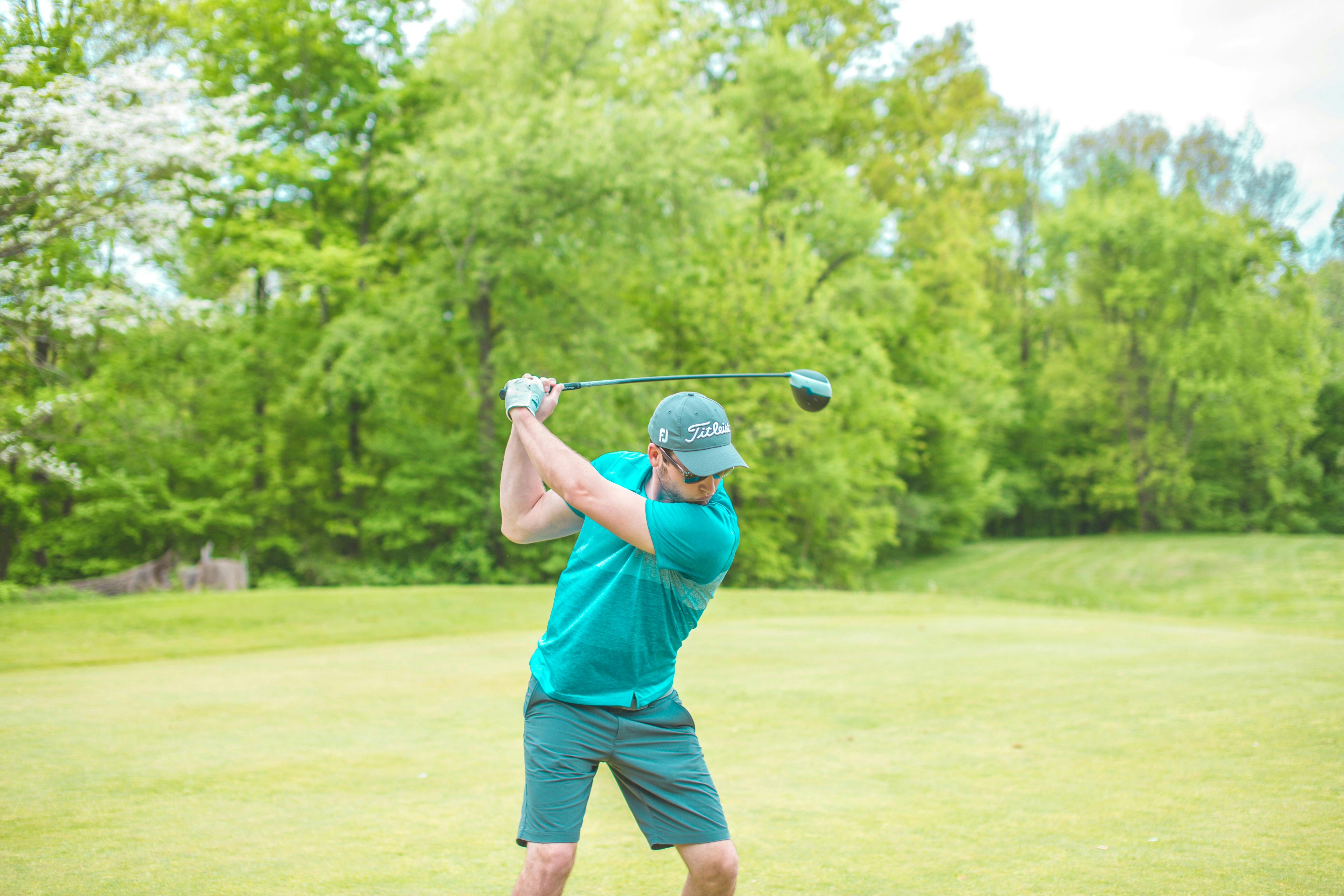 A male golfer in a teal shirt and grey hat is captured mid-swing on a bright green fairway, his club a blur of motion. A lush, dense line of green trees forms the background.