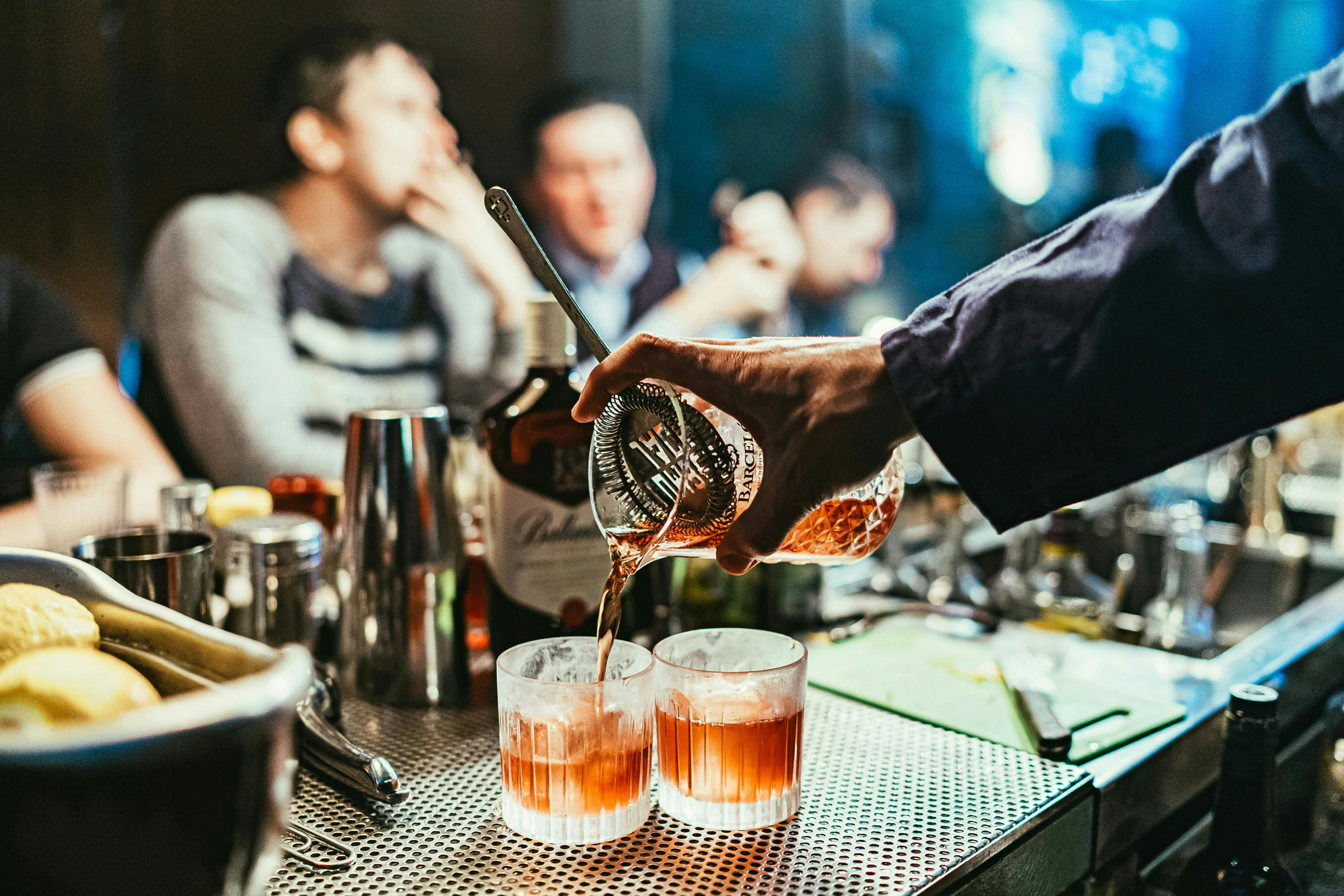 A bartender's hand in a dark blue shirt pours an amber-colored cocktail from a mixing glass with a strainer into two old-fashioned glasses on a metal-grate bar top. The background is dark and out of focus, showing several patrons seated at the bar.
