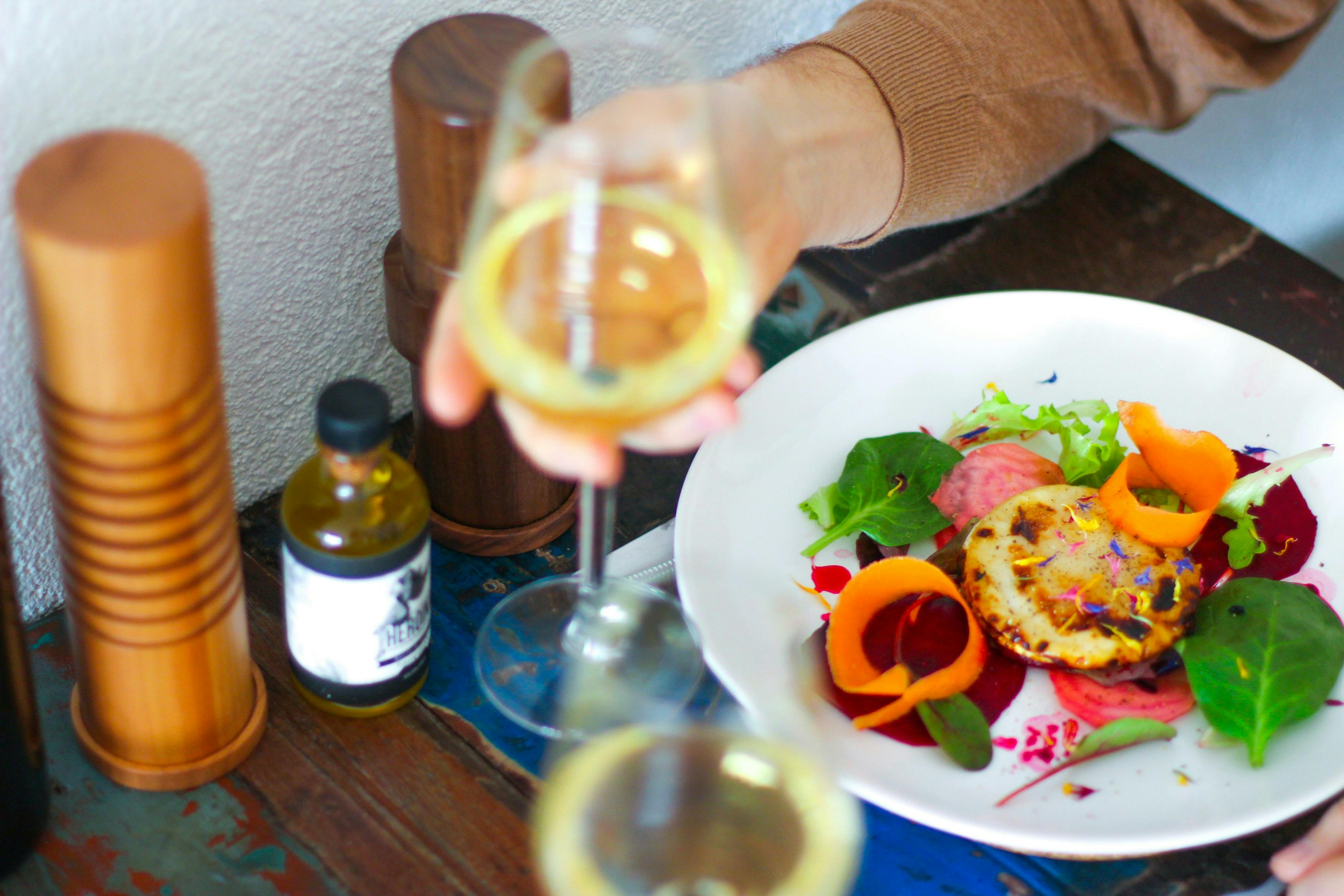 An overhead shot of a hand holding up a glass of white wine over a plate of beautifully plated food. The dish features a seared circular item, possibly cheese or a scallop, garnished with shredded carrots, spinach, and beet slices. A small bottle of olive oil and wooden spice containers sit on the rustic table next to the plate.
