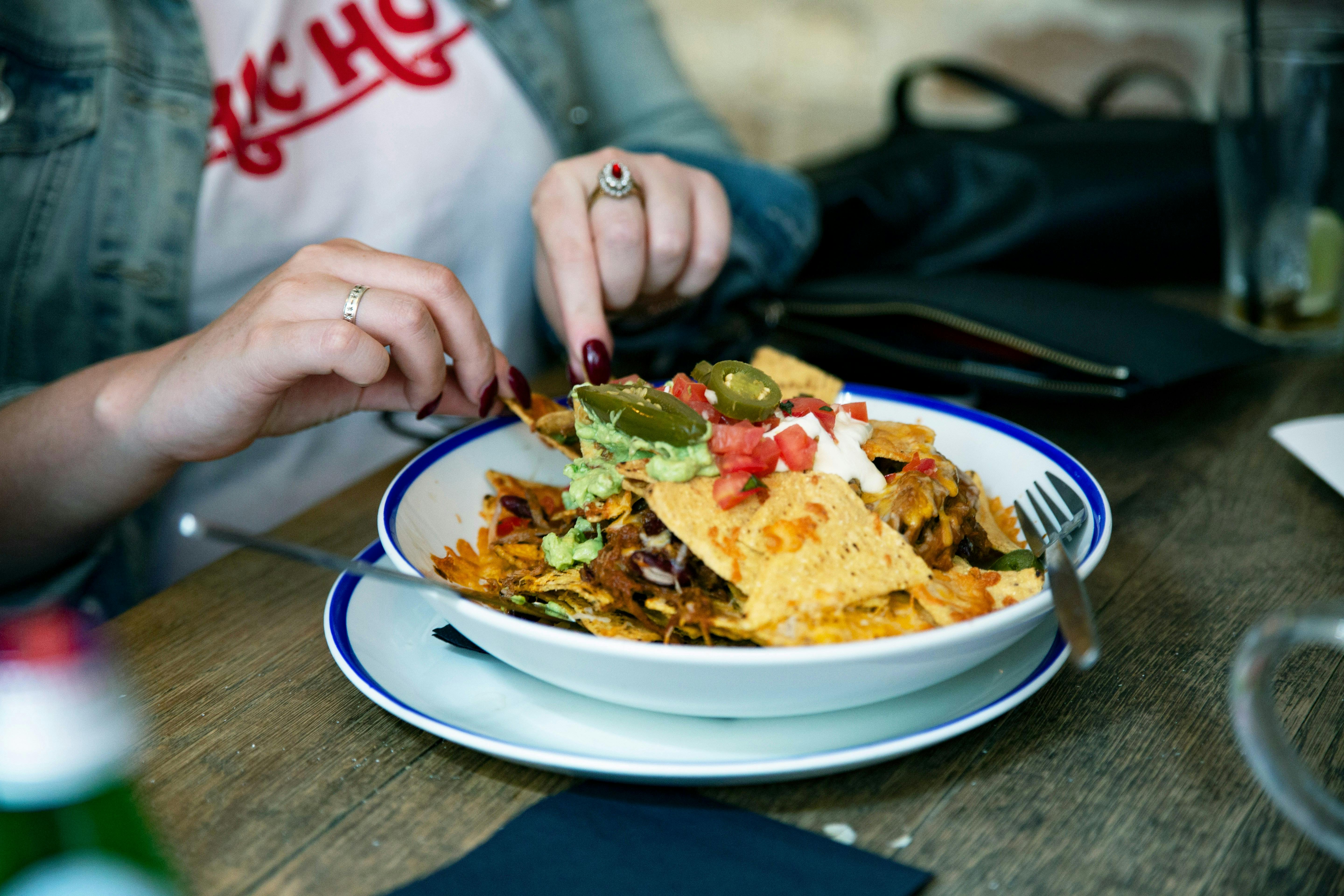A person with dark nail polish is reaching for a tortilla chip from a large, overflowing bowl of nachos. The nachos are topped with melted cheese, guacamole, sour cream, diced tomatoes, and jalapenos. The person is wearing a denim jacket and a white t-shirt.