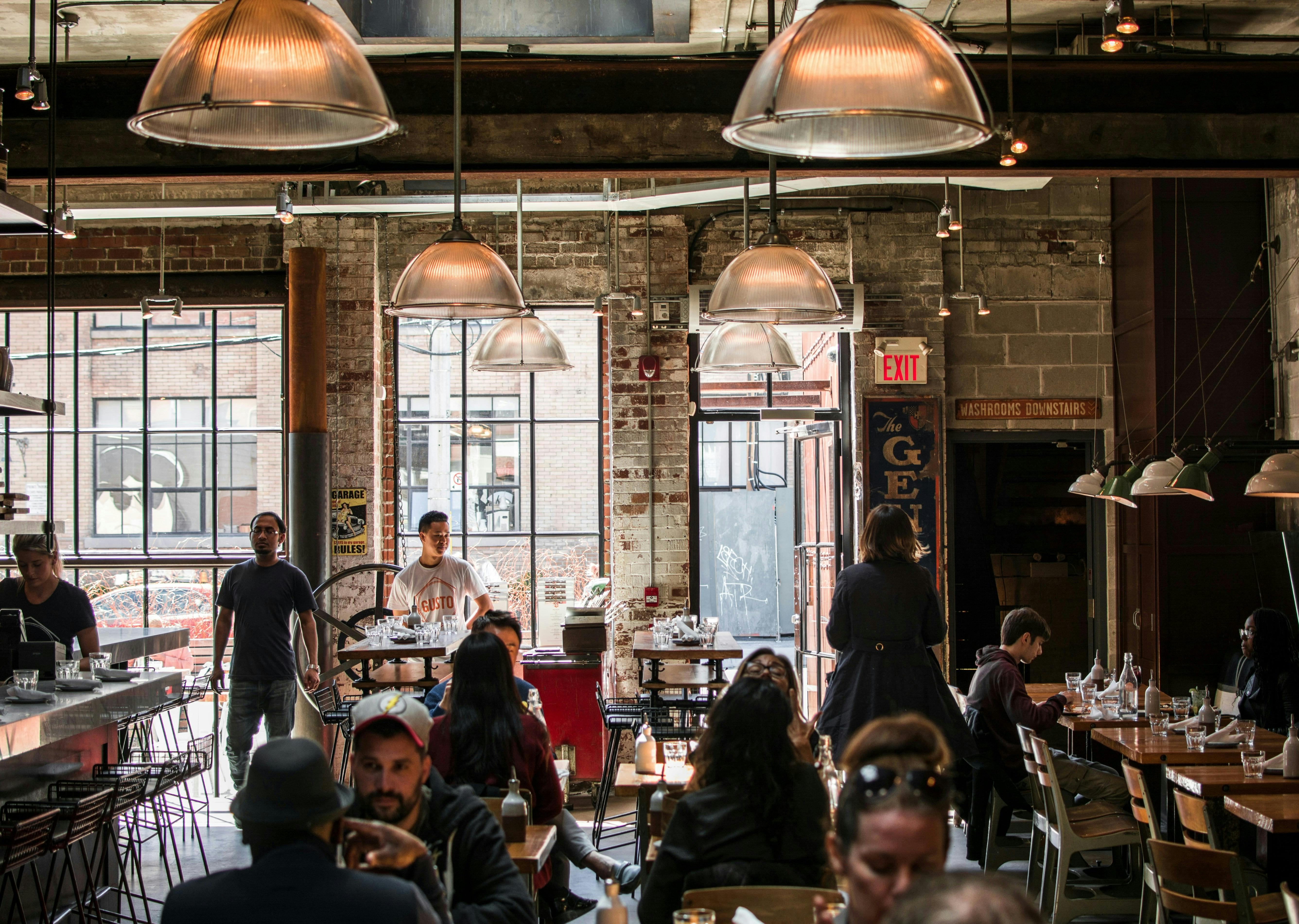 A lively, industrial-style restaurant interior. The space features exposed brick walls, large windows, and overhead metallic pendant lights. Many patrons are seated at wooden tables and a counter. Two people are standing near a doorway in the background.