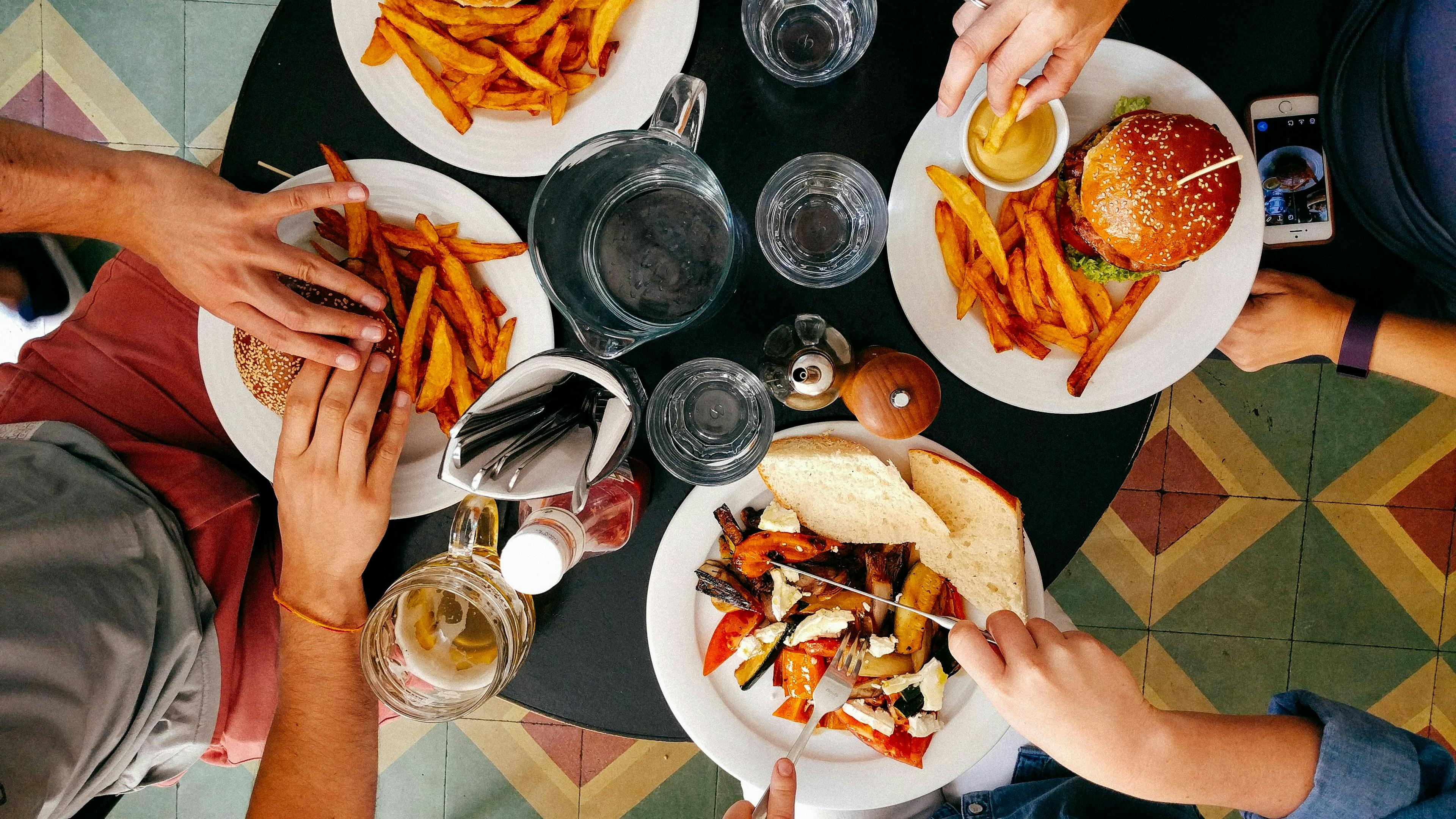 A top-down view of a round table with four people dining. Two plates hold a hamburger and a large serving of french fries. The third plate has a large portion of french fries, and the fourth plate contains grilled vegetables and sliced bread. Hands are reaching for the food, and a pitcher of water and several glasses are on the table.