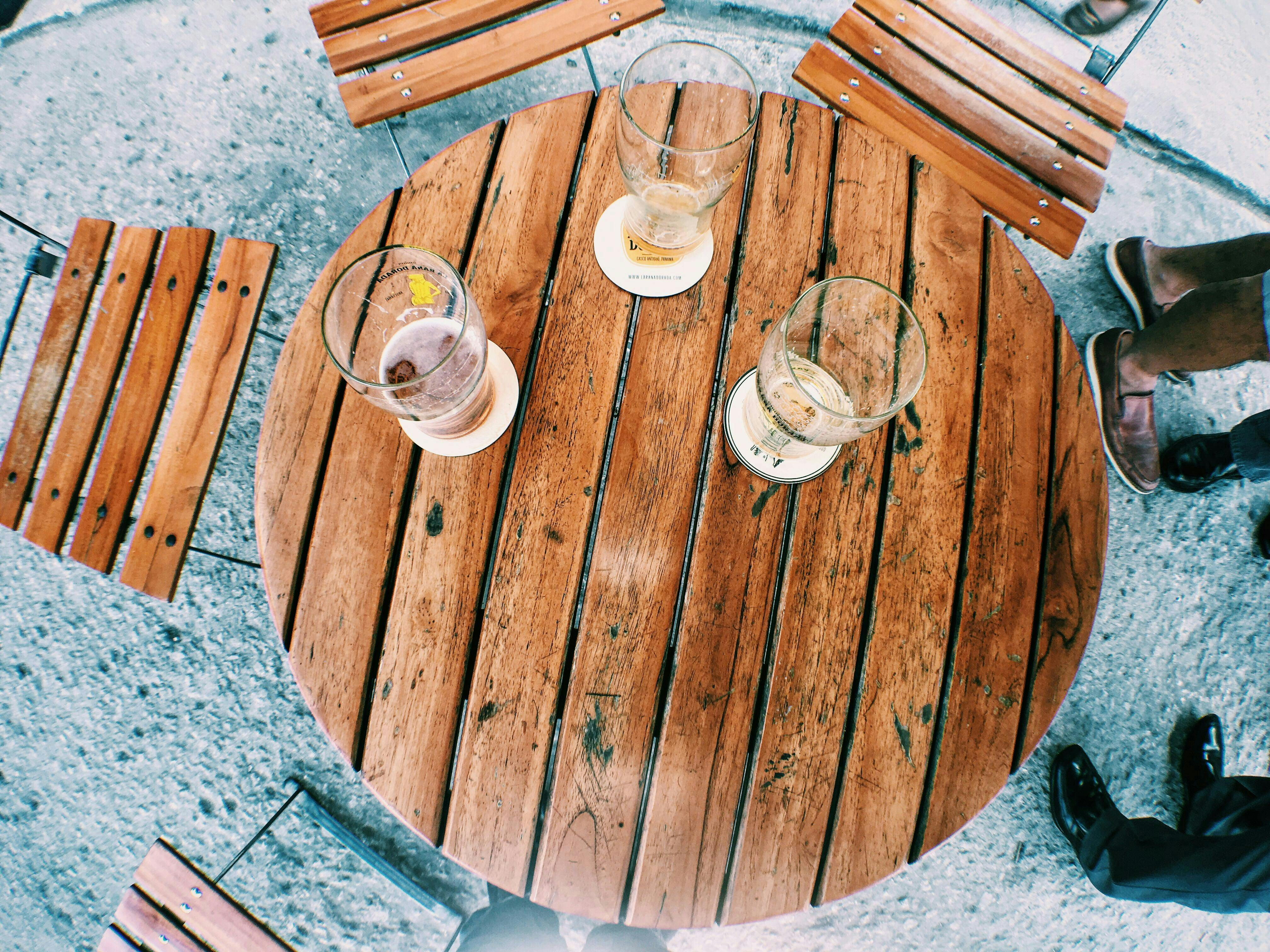 A top-down, high-angle view of a round, slatted wooden table outdoors with three partially empty glasses of beer on coasters. Three wooden chairs are pulled away from the table, and the feet of two people wearing brown and black shoes are visible on the grey concrete ground.