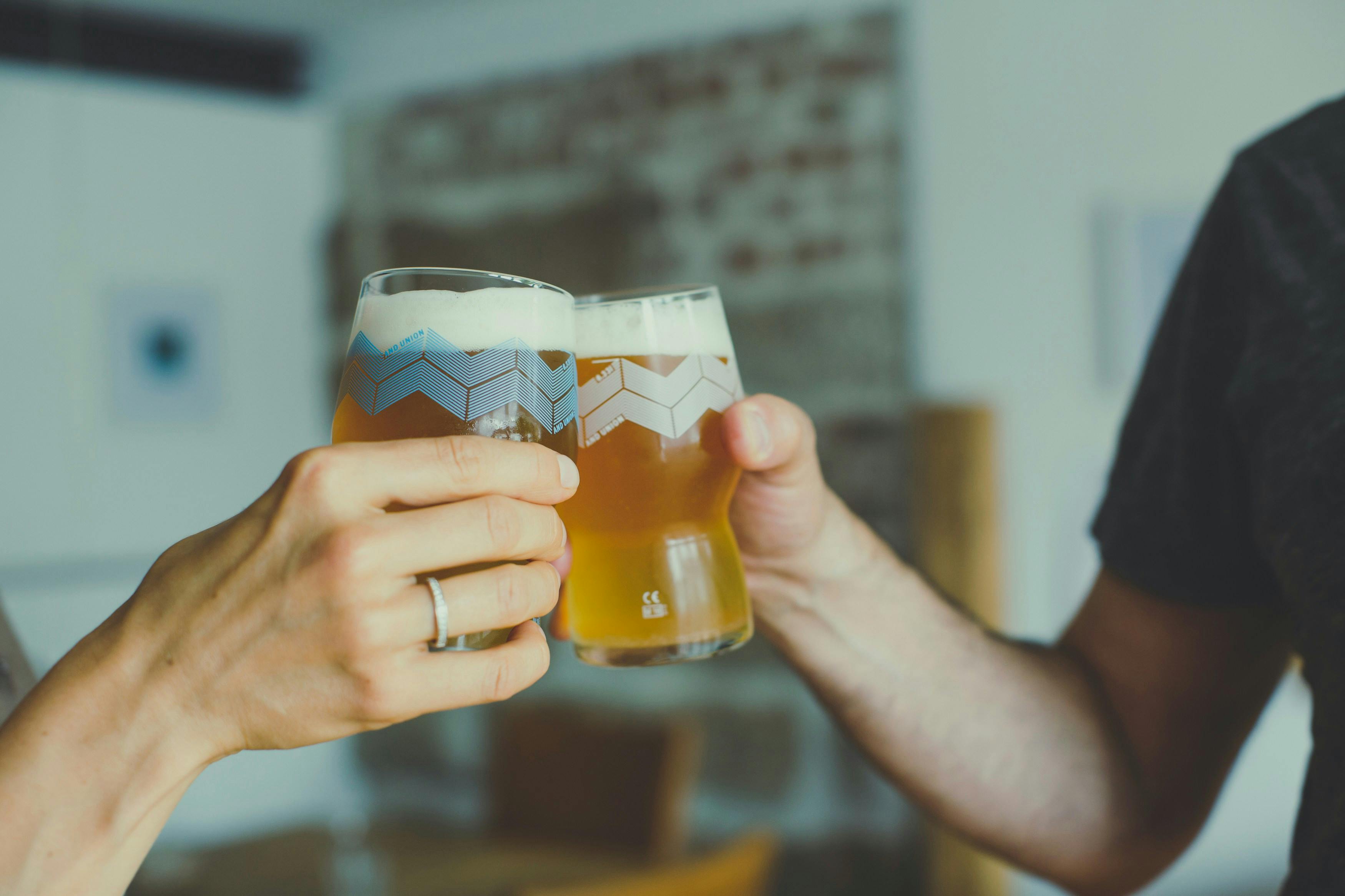 A close-up, focused shot of two people cheersing with glasses of beer. The glasses are unique, with a light blue and white geometric pattern on the upper half. A woman's hand, wearing a ring, is on the left, and a man's arm and hand are on the right.