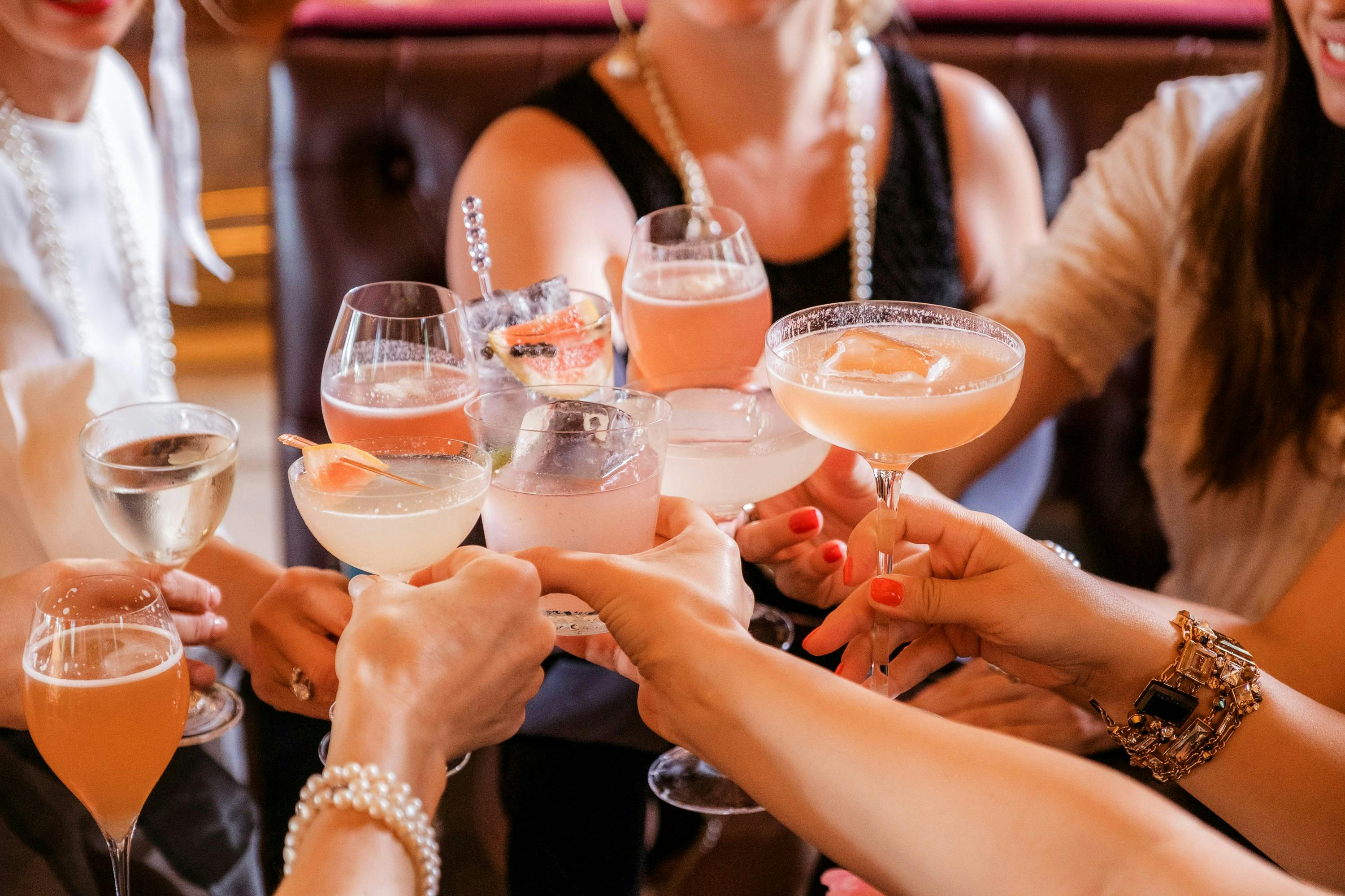 A close-up shot of a group of elegantly dressed people raising various cocktails and glasses of sparkling wine in a celebratory cheers. The drinks are pink, white, and orange, and the women's hands are decorated with pearl bracelets and gold cuffs.