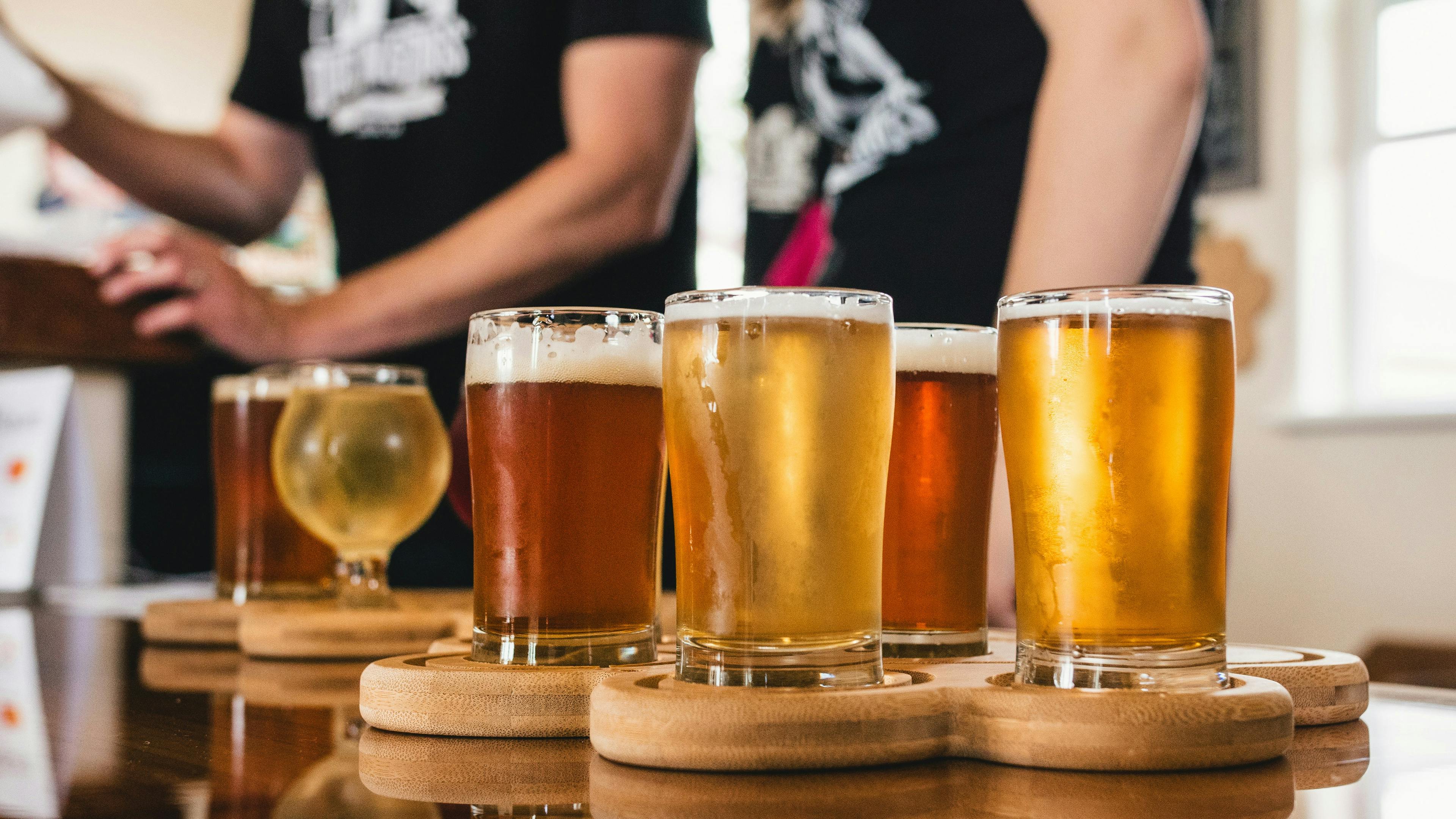 A line of six glasses of various beers—including pale, amber, and dark varieties—are arranged on small wooden serving paddles on a reflective wooden bar top. Two blurry figures in black t-shirts are visible standing in the background, suggesting a brewery or taproom setting.
