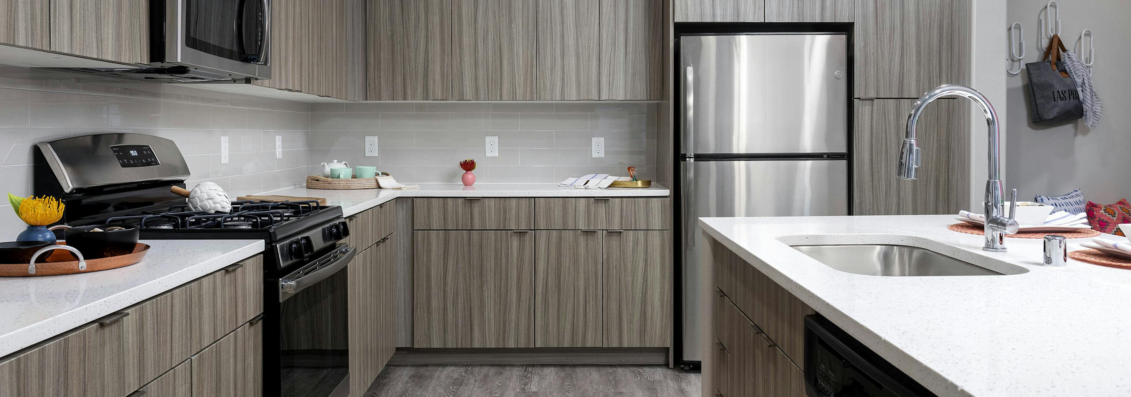 Interior view of AMLI Camarillo apartment kitchen with light wood grain cabinets, stainless appliances, quartz counters and tile backsplash