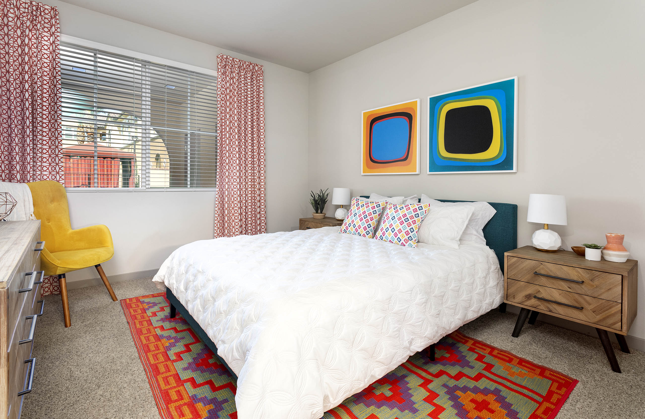 Interior view of AMLI Camarillo apartment bedroom with white bedding, colorful area rug, yellow chair and abstract wall art.