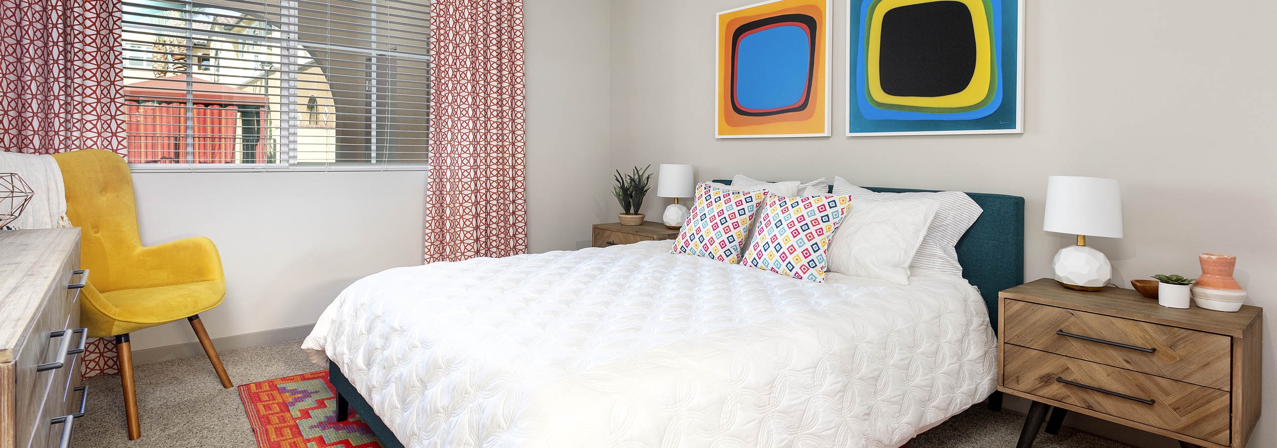 Interior view of AMLI Camarillo apartment bedroom with white bedding, colorful area rug, yellow chair and abstract wall art.