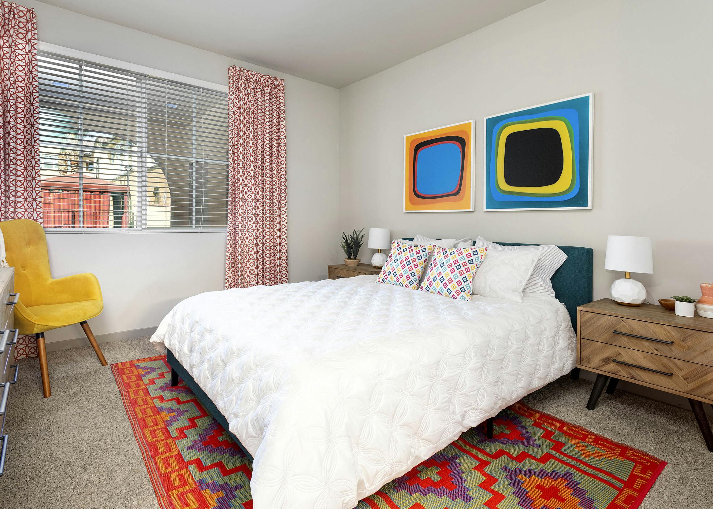 Interior view of AMLI Camarillo apartment bedroom with white bedding, colorful area rug, yellow chair and abstract wall art.