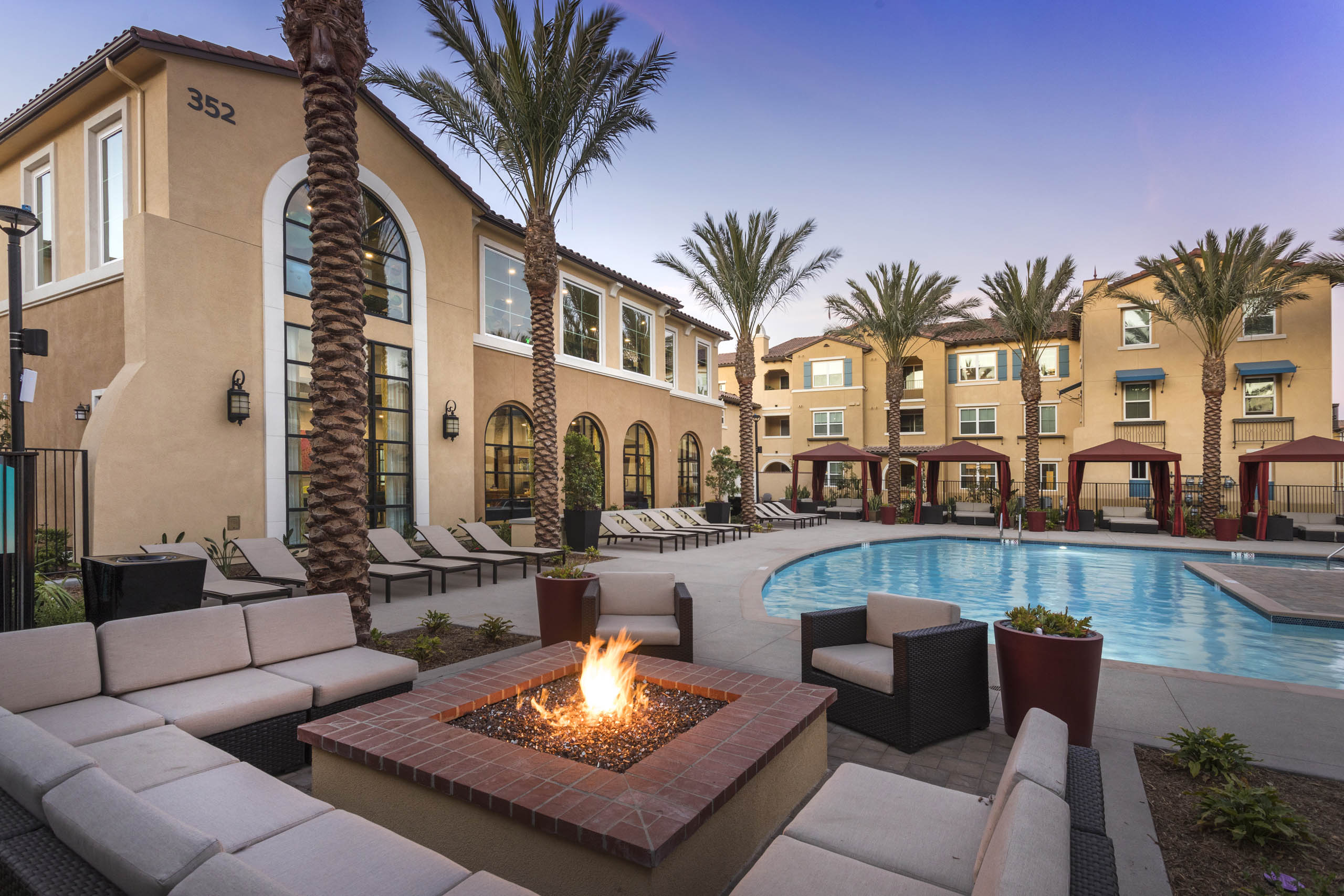 Poolside firepit lounge at AMLI Camarillo apartments with sectional cushioned seating, tropical landscaping, pool and gold building in background.