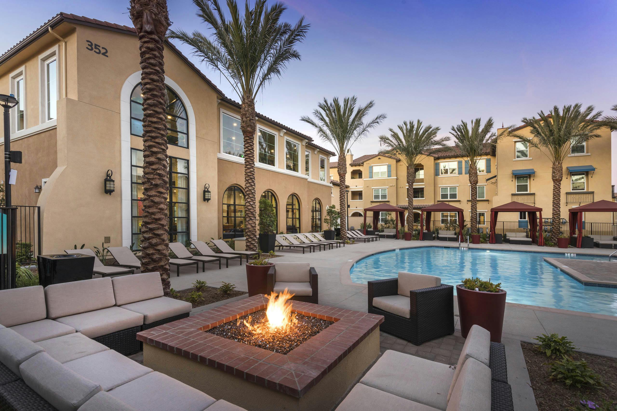Poolside firepit lounge at AMLI Camarillo apartments with sectional cushioned seating, tropical landscaping, pool and gold building in background.