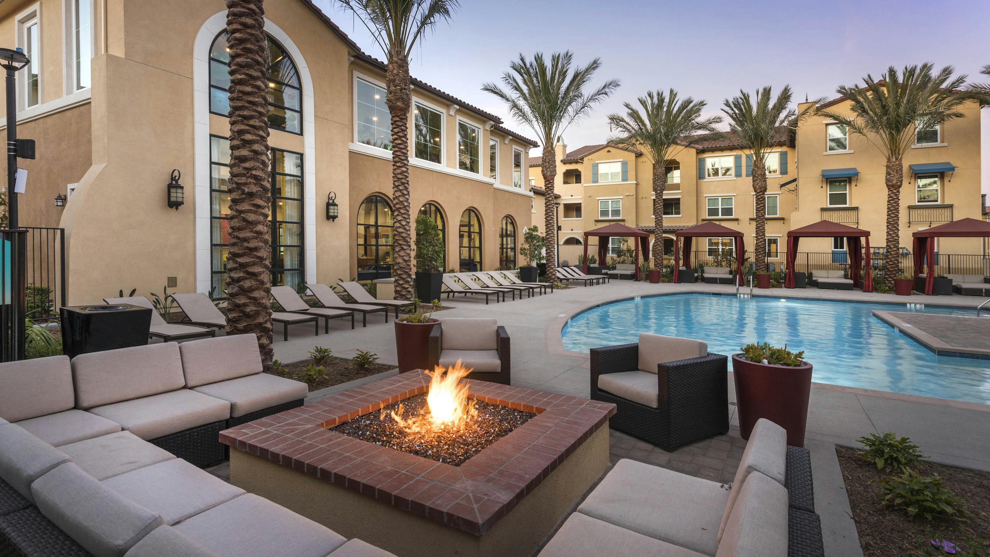 Poolside firepit lounge at AMLI Camarillo apartments with sectional cushioned seating, tropical landscaping, pool and gold building in background.
