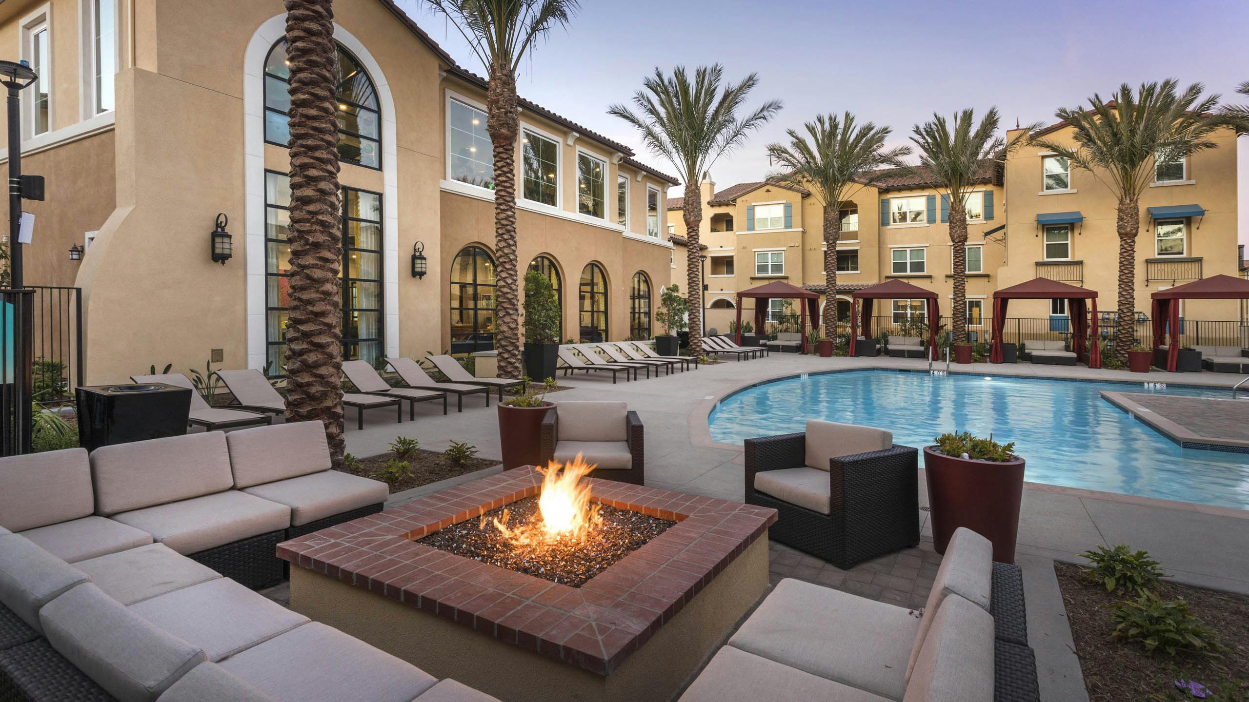 Poolside firepit lounge at AMLI Camarillo apartments with sectional cushioned seating, tropical landscaping, pool and gold building in background.