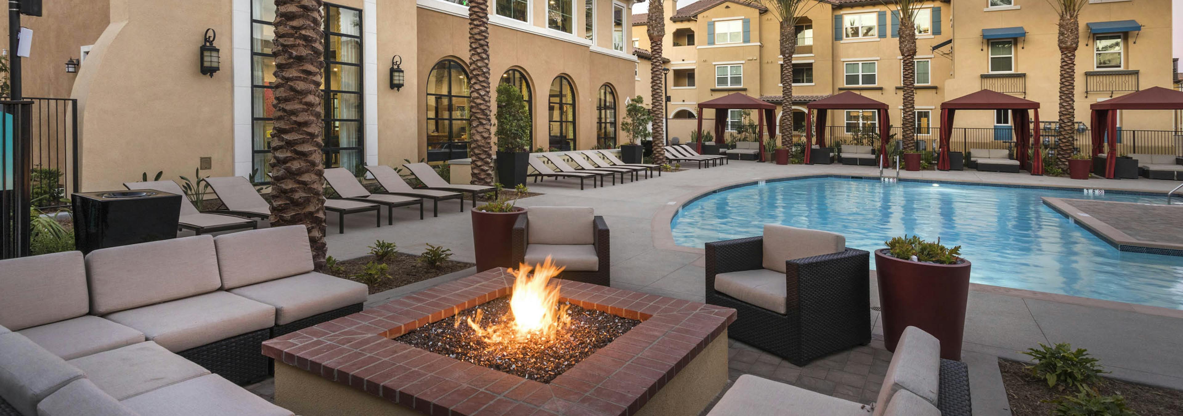 Poolside firepit lounge at AMLI Camarillo apartments with sectional cushioned seating, tropical landscaping, pool and gold building in background.