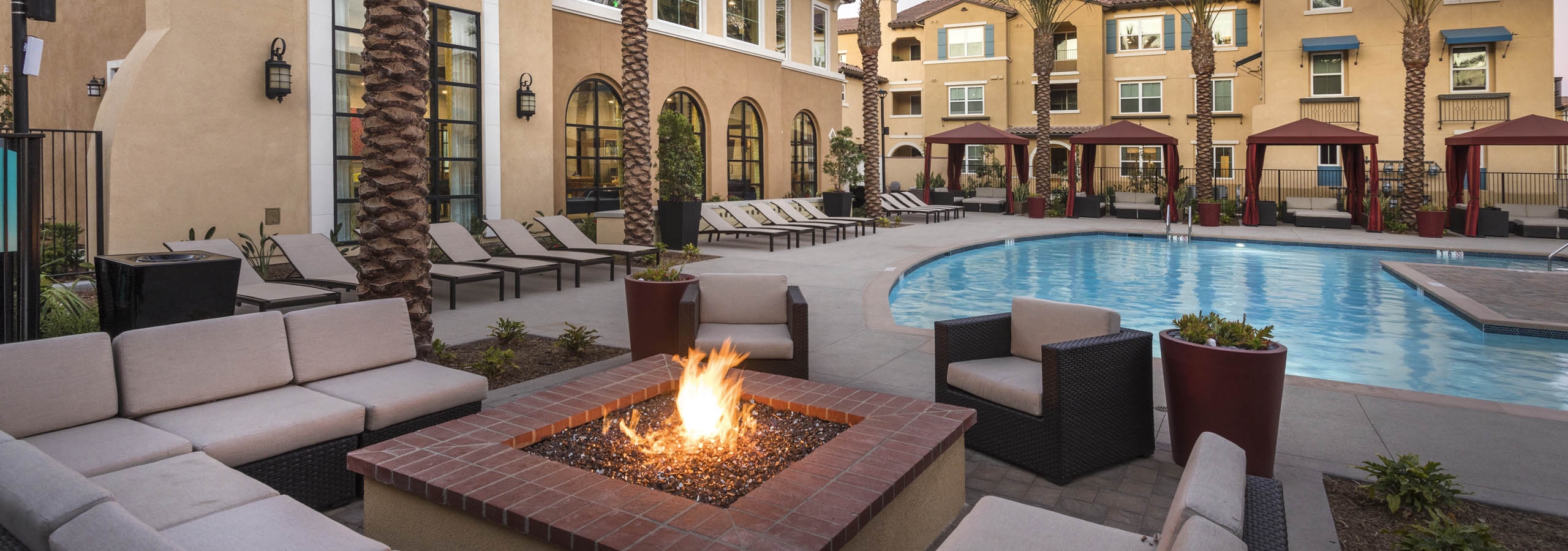 Poolside firepit lounge at AMLI Camarillo apartments with sectional cushioned seating, tropical landscaping, pool and gold building in background.
