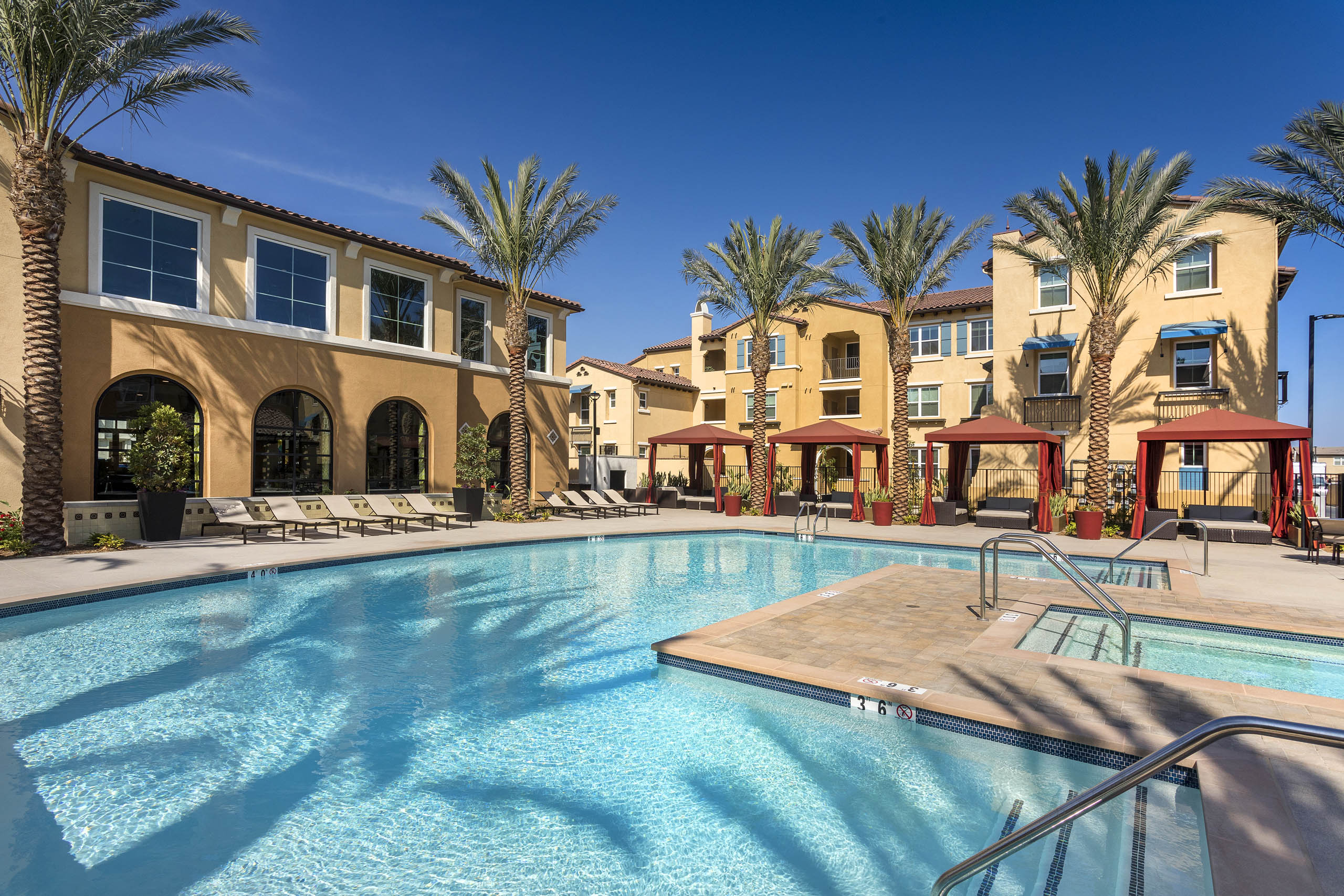 Daytime poolside view at AMLI Camarillo apartments with spa, red cabanas, lounge chairs and tan buildings surrounded by palm trees