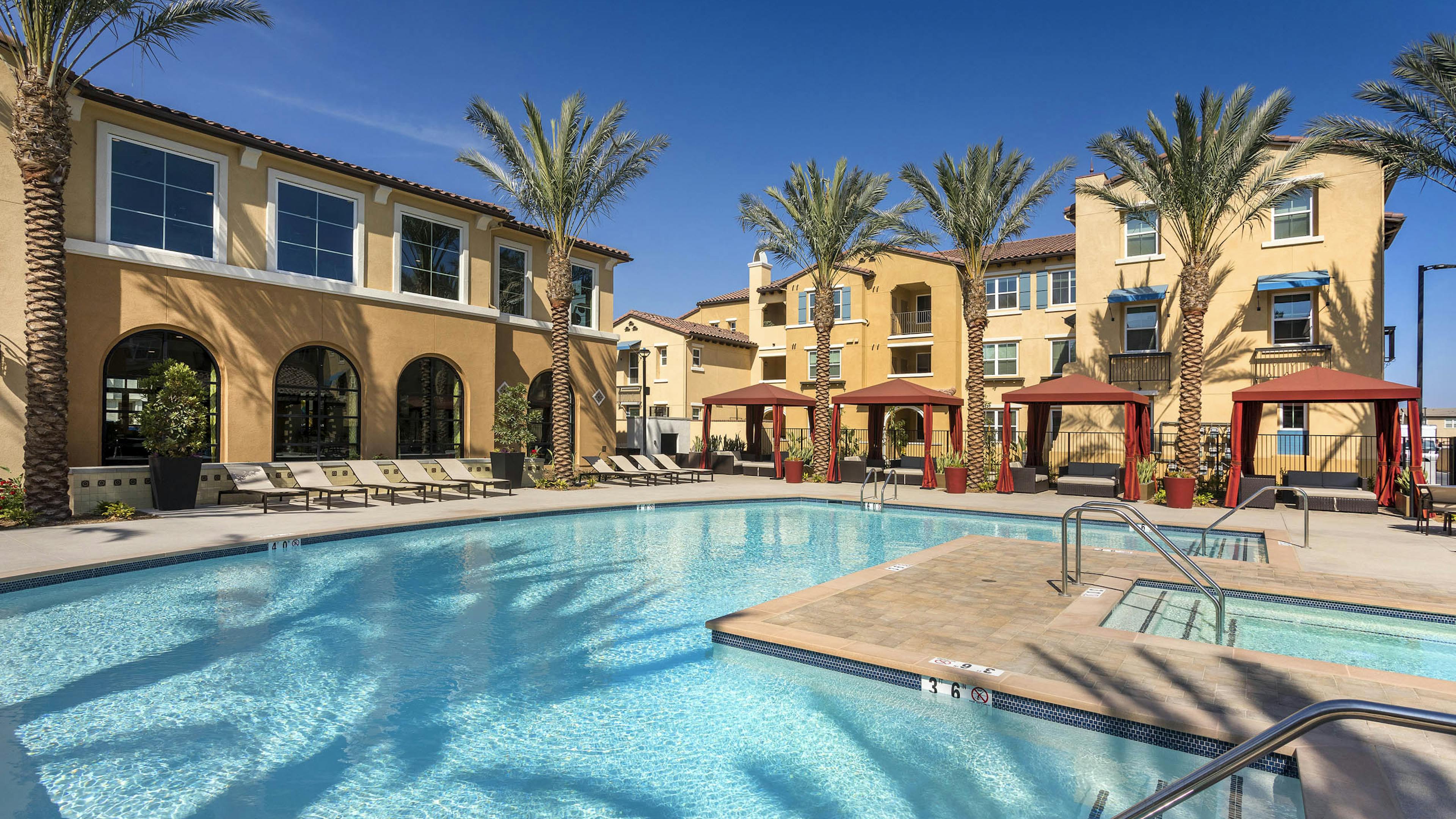 Daytime poolside view at AMLI Camarillo apartments with spa, red cabanas, lounge chairs and tan buildings surrounded by palm trees