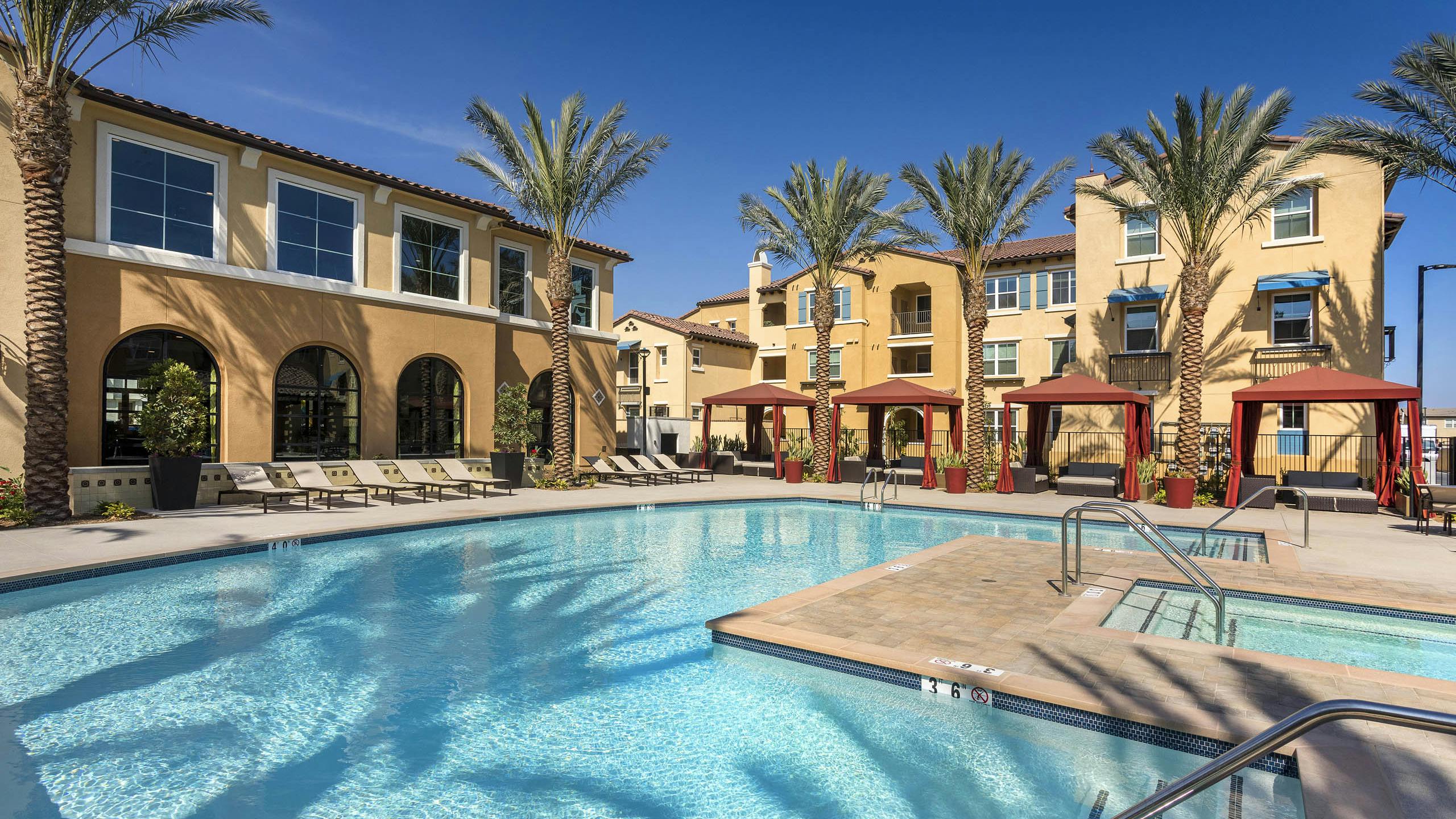 Daytime poolside view at AMLI Camarillo apartments with spa, red cabanas, lounge chairs and tan buildings surrounded by palm trees