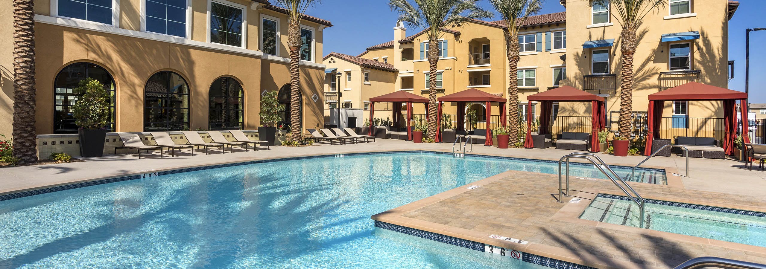 Daytime poolside view at AMLI Camarillo apartments with spa, red cabanas, lounge chairs and tan buildings surrounded by palm trees