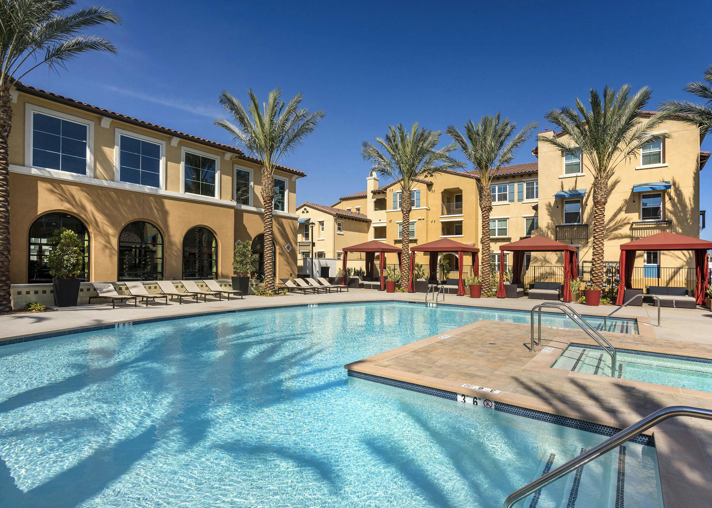 Daytime poolside view at AMLI Camarillo apartments with spa, red cabanas, lounge chairs and tan buildings surrounded by palm trees
