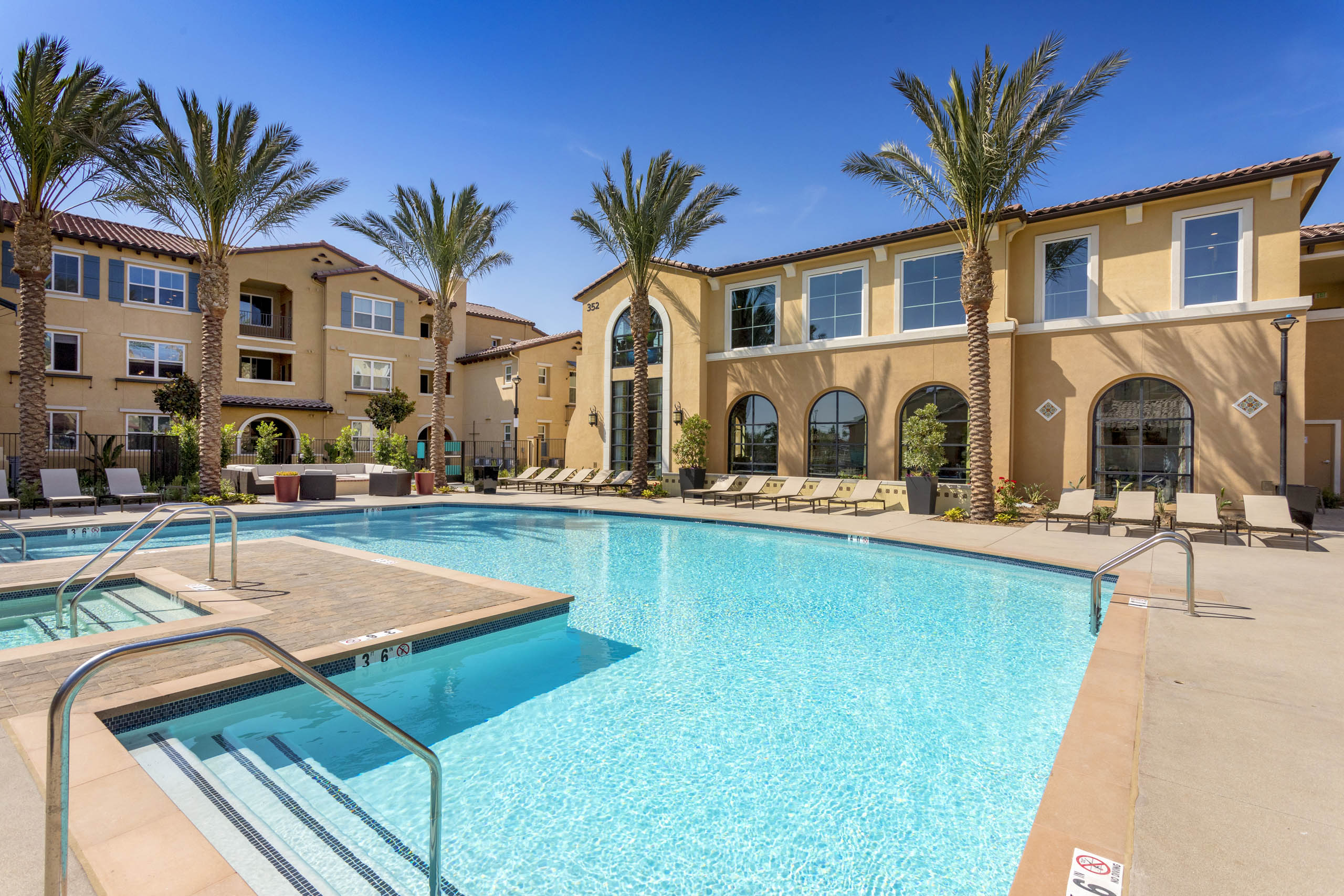 Daytime view of AMLI Camarillo apartments bright blue pool lined with tan loungers, tall palm trees, and tan stucco buildings. 