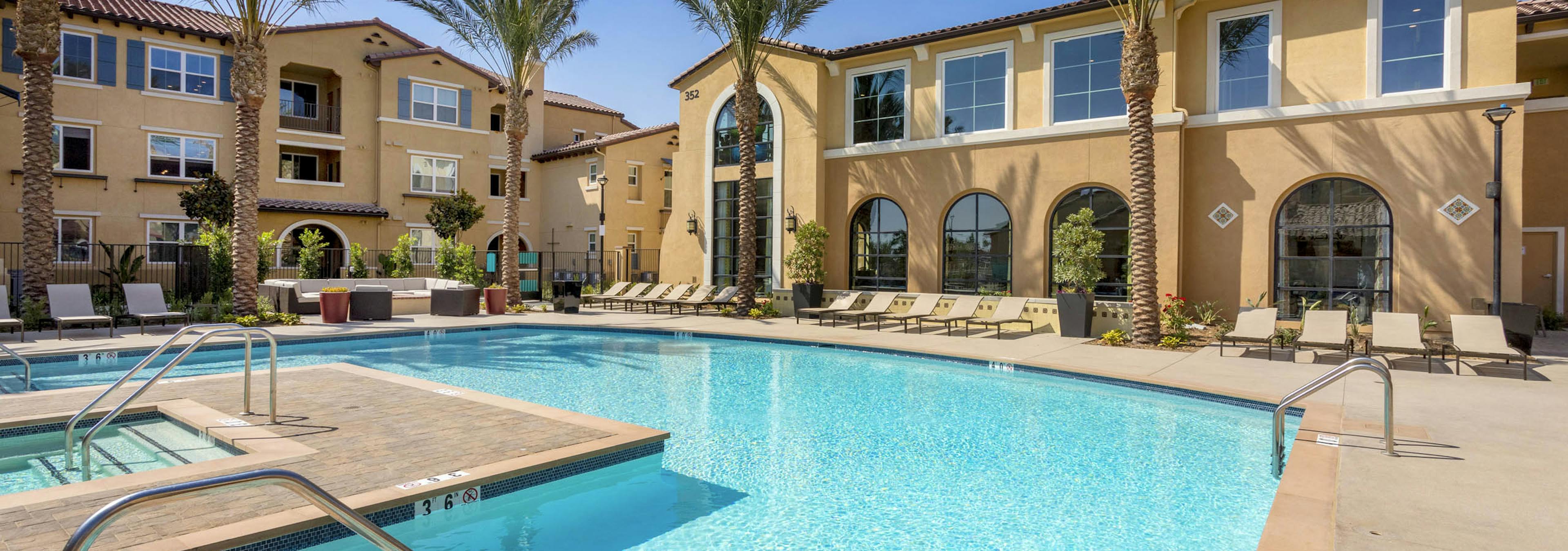 Daytime view of AMLI Camarillo apartments bright blue pool lined with tan loungers, tall palm trees, and tan stucco buildings. 