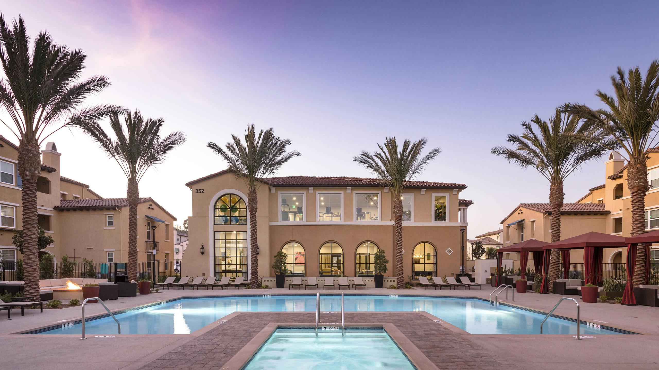 Dusk view of AMLI Camarillo apartments featuring a central hot tub, glowing pool, red cabanas and palm-lined buildings