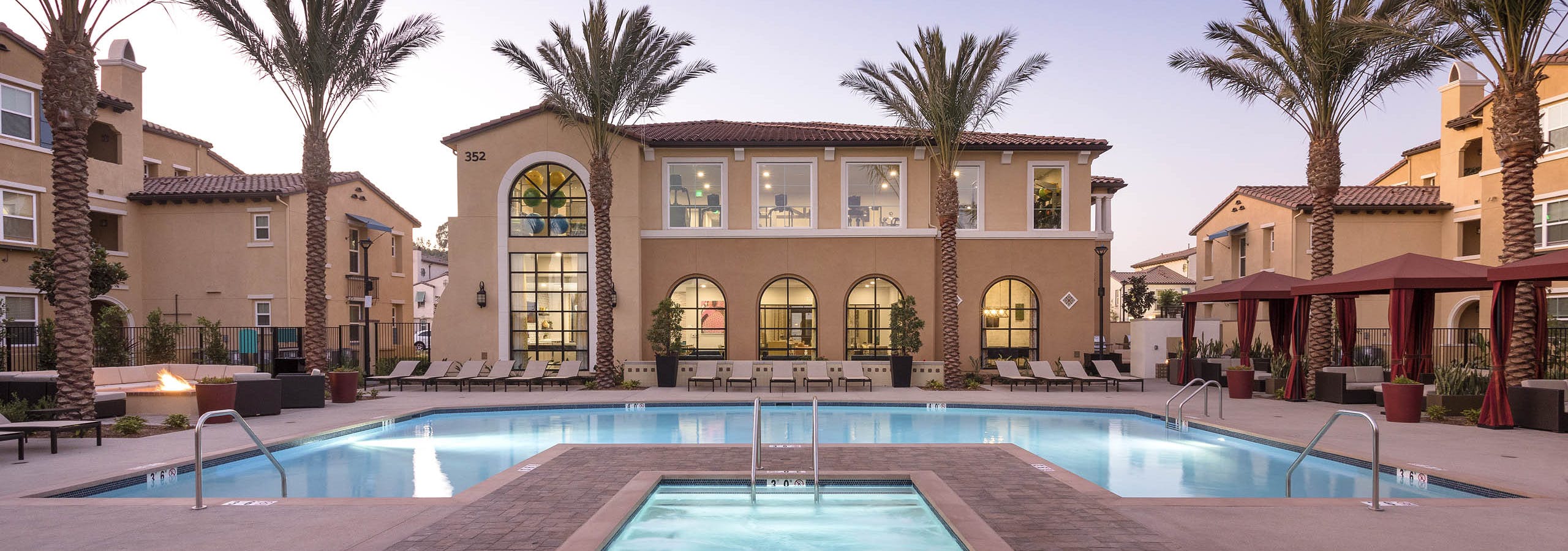 Dusk view of AMLI Camarillo apartments featuring a central hot tub, glowing pool, red cabanas and palm-lined buildings