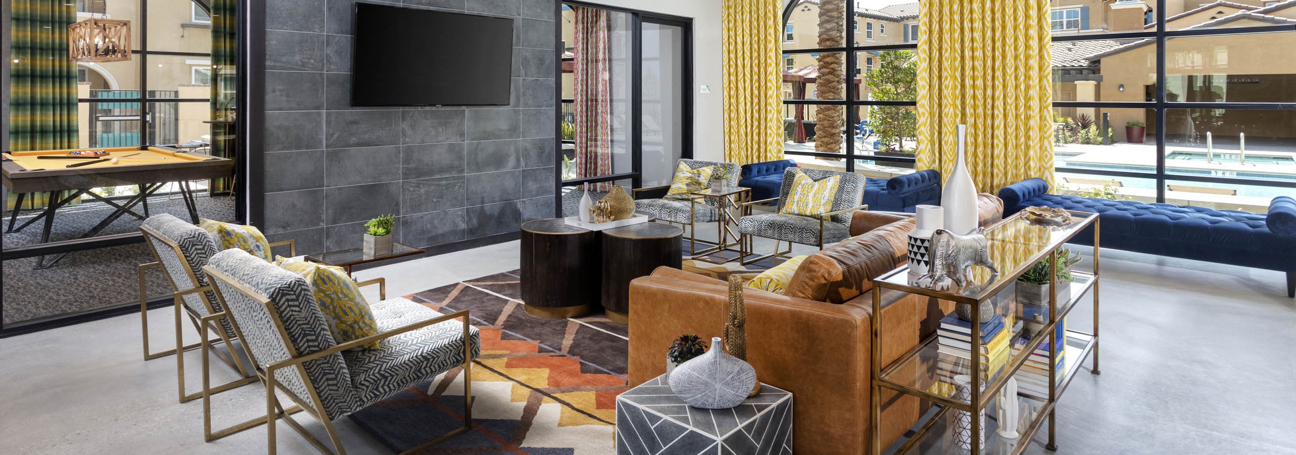 Interior view of AMLI Camarillo clubhouse lounge with bold decor, leather seating, mounted TV, and large windows overlooking the pool