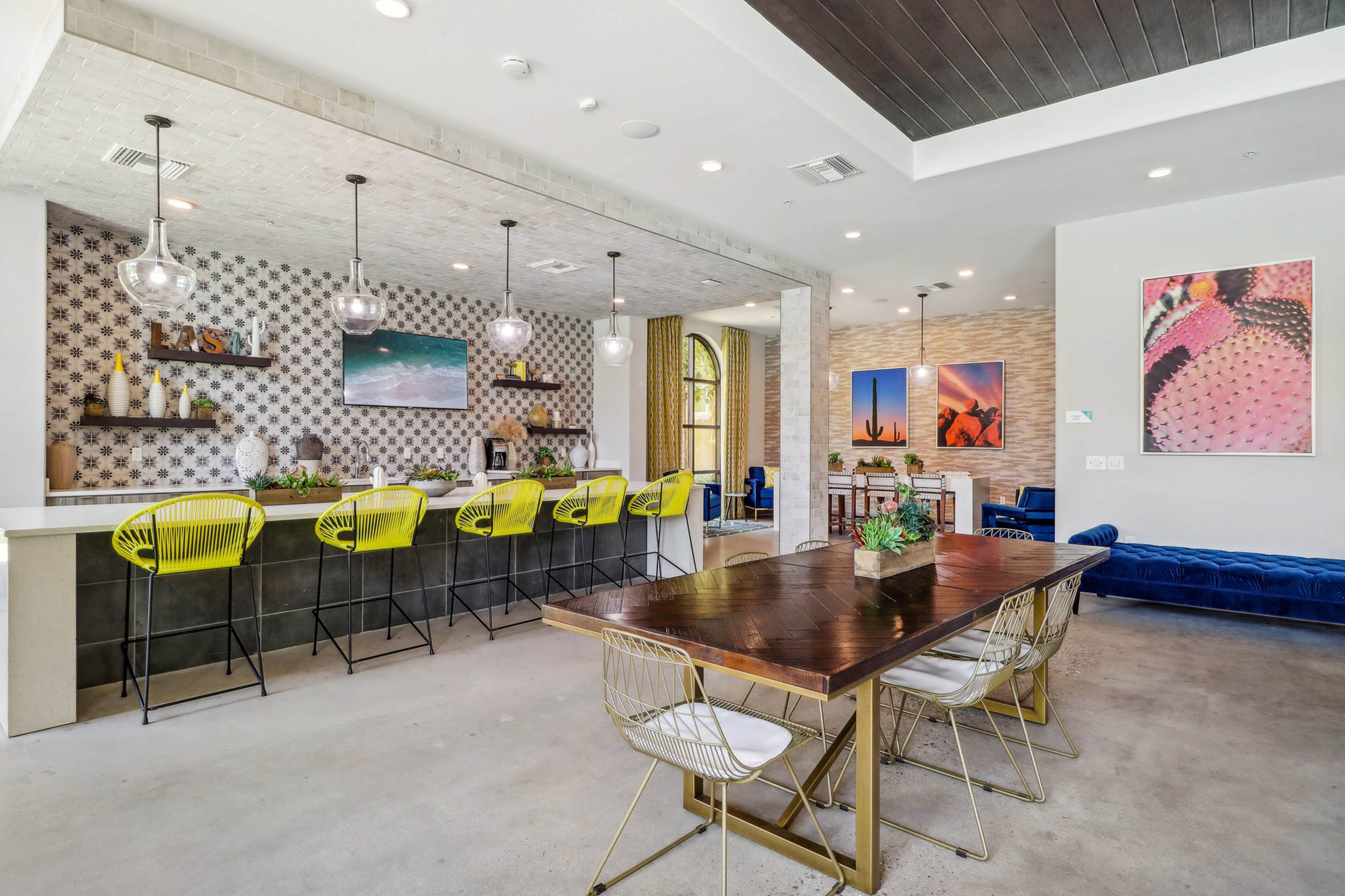 Clubhouse interior at AMLI Camarillo apartments with dining table, yellow barstools, cactus wall art, and pendant lights over a tiled bar