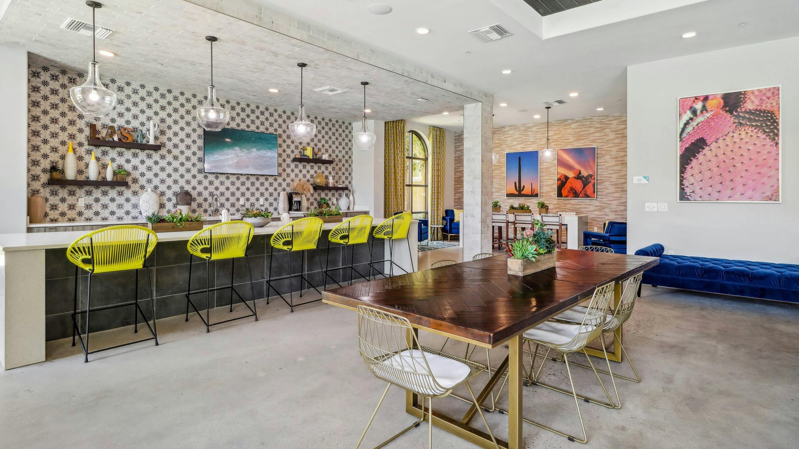 Clubhouse interior at AMLI Camarillo apartments with dining table, yellow barstools, cactus wall art, and pendant lights over a tiled bar