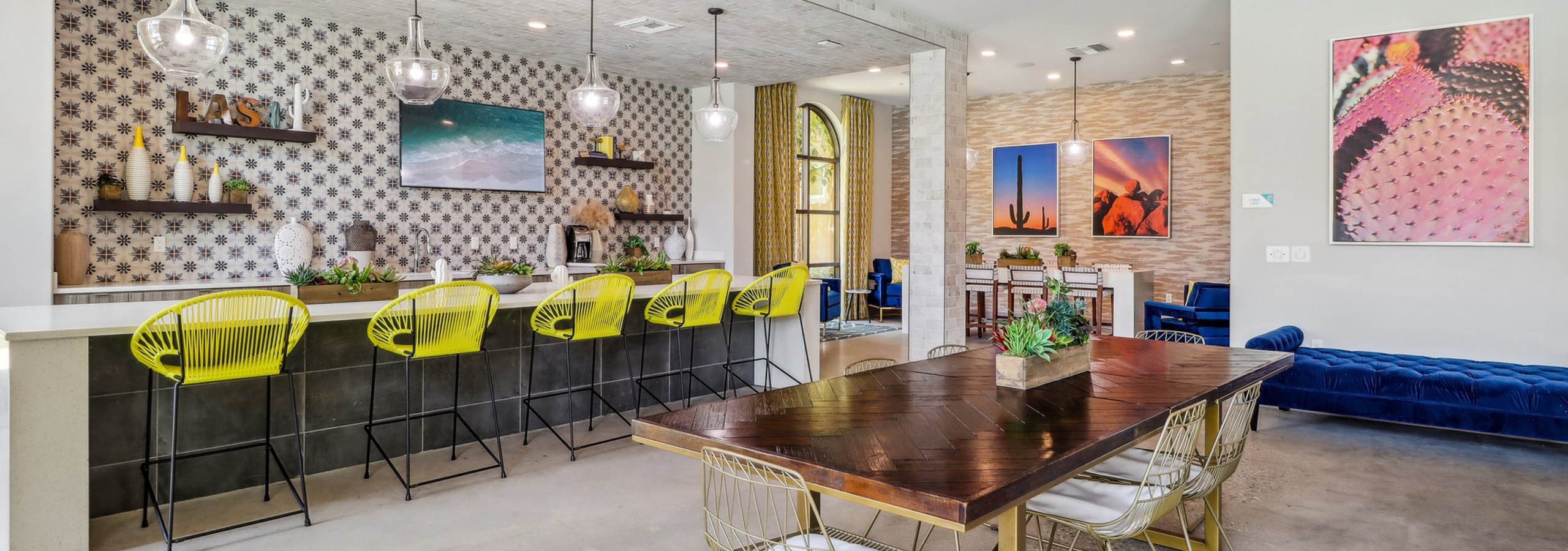 Clubhouse interior at AMLI Camarillo apartments with dining table, yellow barstools, cactus wall art, and pendant lights over a tiled bar