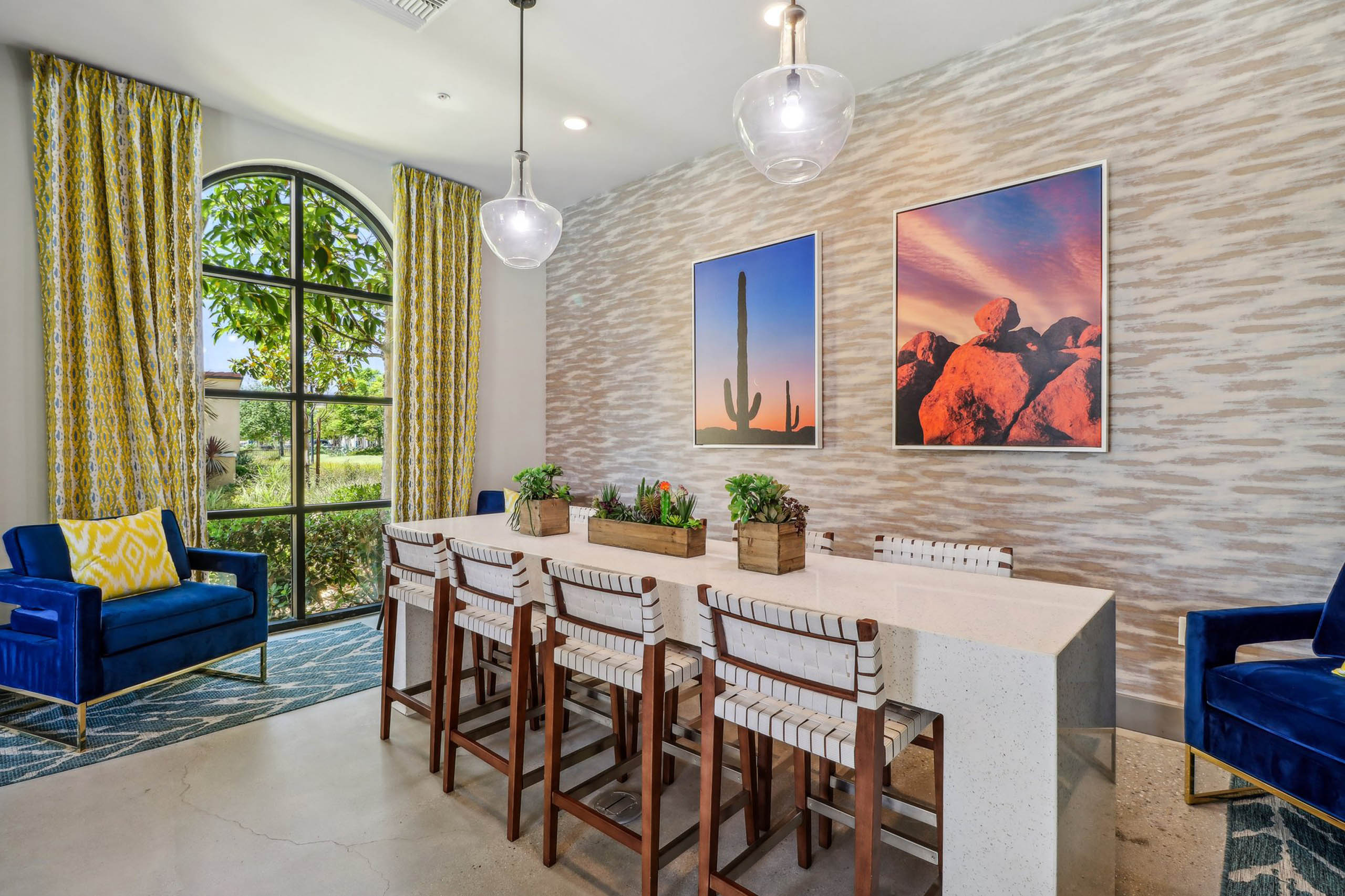 Clubroom at AMLI Camarillo apartments featuring a quartz-topped table, wood chairs, desert artwork and arched window with yellow drapes